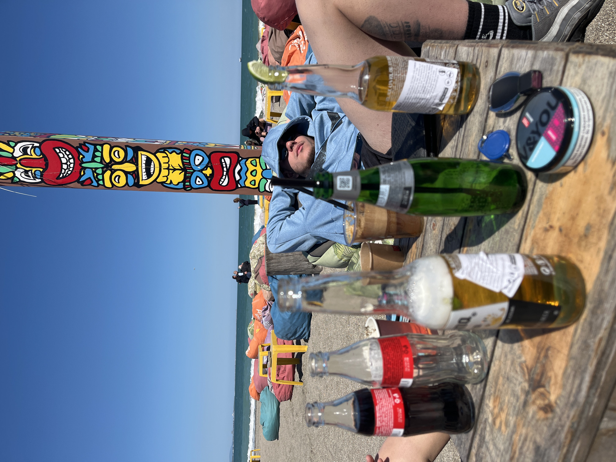 The image captures a scene at the beach in Burgas, Bulgaria. A man wearing a blue hoodie and sunglasses reclines on a beach chair with a wooden table to his side. Several empty beer and cola bottles lie on the table, along with a can of snus and keys. A colorful totem pole stands nearby. The beach is populated with people and beanbag chairs. The sky is clear and blue, and the sea is visible in the background.