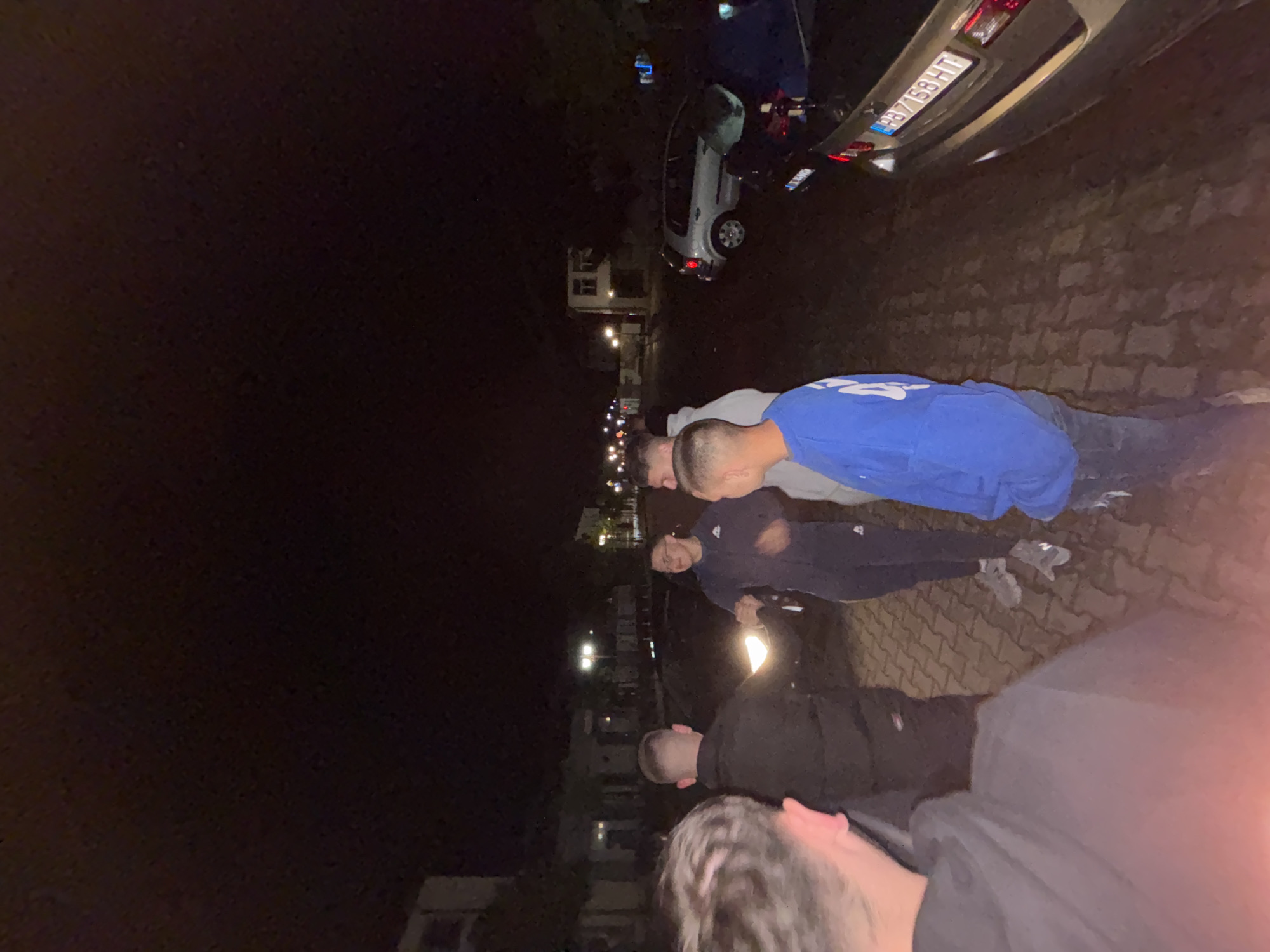 At night in Zlatograd, Bulgaria, a group of approximately five people stands near a cobblestone road. A car is parked nearby, bearing a license plate reading "AB8758HT". Streetlights illuminate the area, with buildings visible in the background. The people seem to be engaging in a conversation. One person is wearing a blue shirt and jeans, another is wearing a dark-colored jacket.