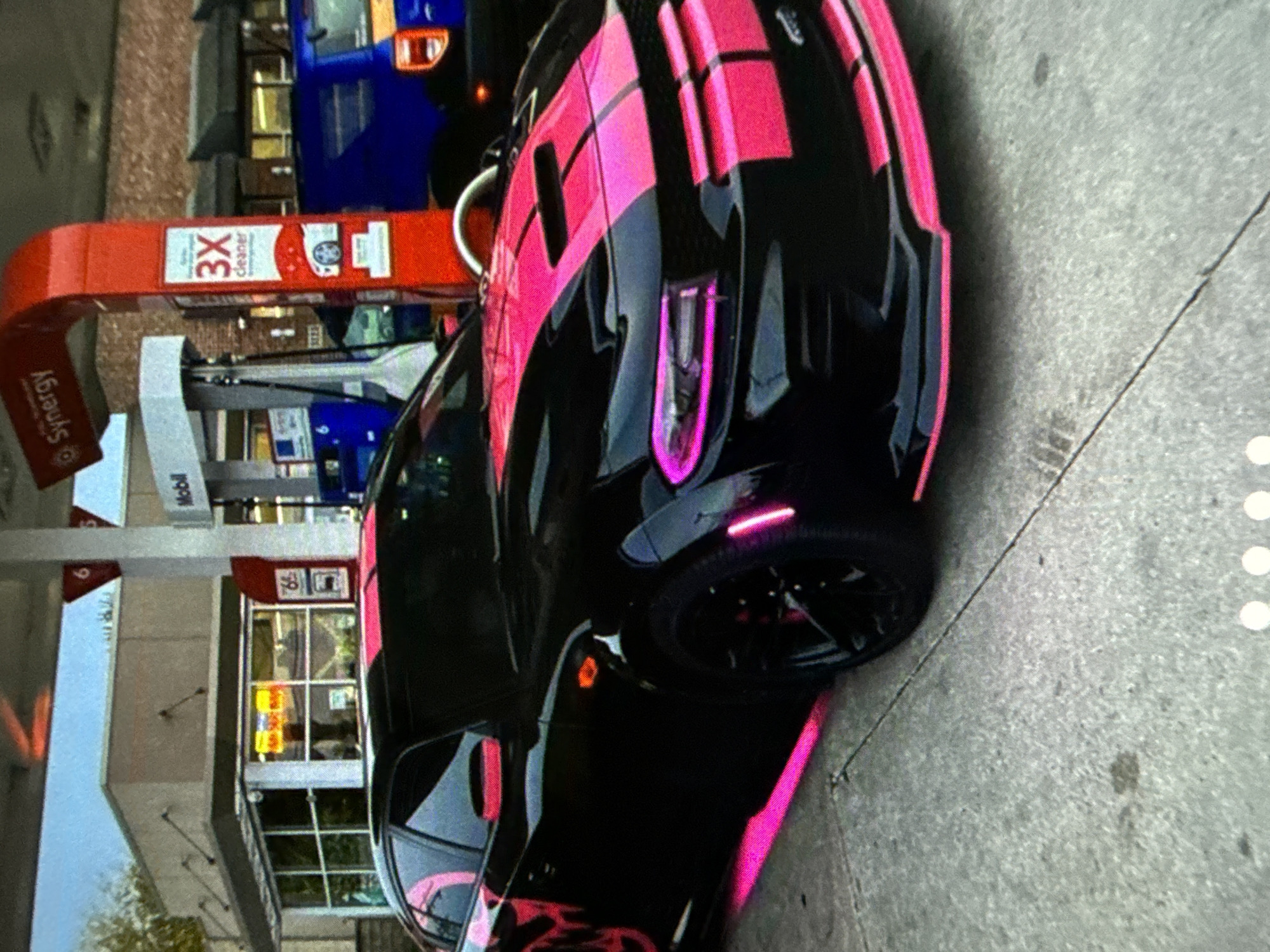 A black car with pink stripes is parked at a Mobil gas station. The gas station has a red canopy with the words "Synergy" and "3X Cleaner" on it. The price of gas is visible as 65. The environment appears to be urban.