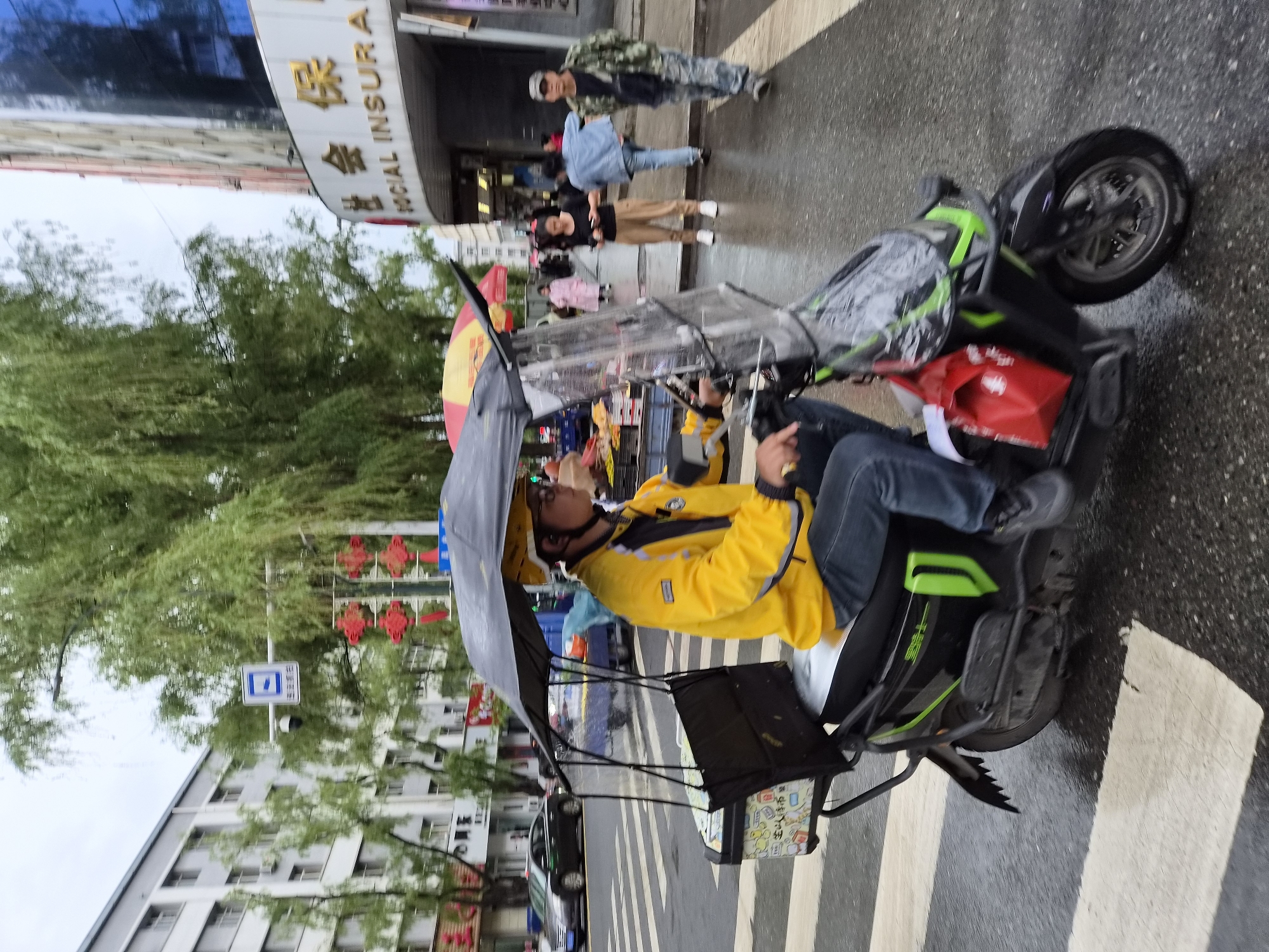 An outdoor, ground-level, medium shot captures a person riding a black and green electric scooter with a canopy and windshield on a wet, paved street, indicated by a crosswalk with white stripes and reflective, wet pavement. The rider, facing right in profile, wears a bright yellow jacket with black and white accents, dark pants, and a yellow helmet. A black box with a colorful graphic design is mounted on the back of the scooter. A red bag is attached to the side of the scooter. In the background, several buildings line the street, one with a curved facade displaying "SOCIAL INSURANCE" in gold lettering on a white background. Trees with green foliage hang over the street. People are visible walking on the sidewalk. A traffic sign with a blue and white symbol is suspended over the road. The overall atmosphere suggests a functional urban environment during overcast or rainy weather.