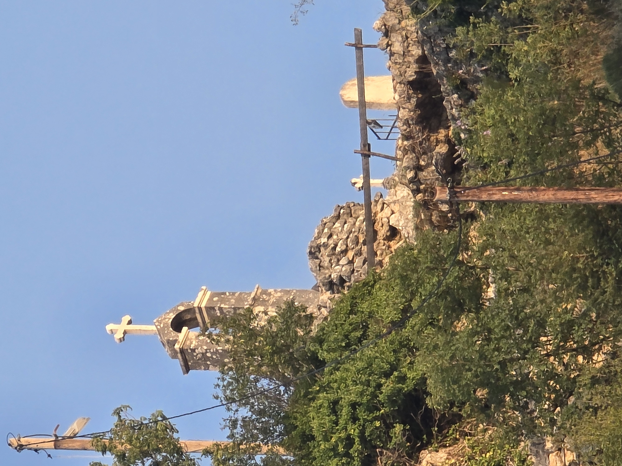 A small, stone bell tower with a cross on top stands on a rocky outcrop in Medun, Montenegro. The tower is part of a ruined structure, with loose stones and vegetation surrounding it. A wooden fence is visible on the outcrop, and a tall wooden pole with wires is in the foreground to the left. The sky is clear and blue.