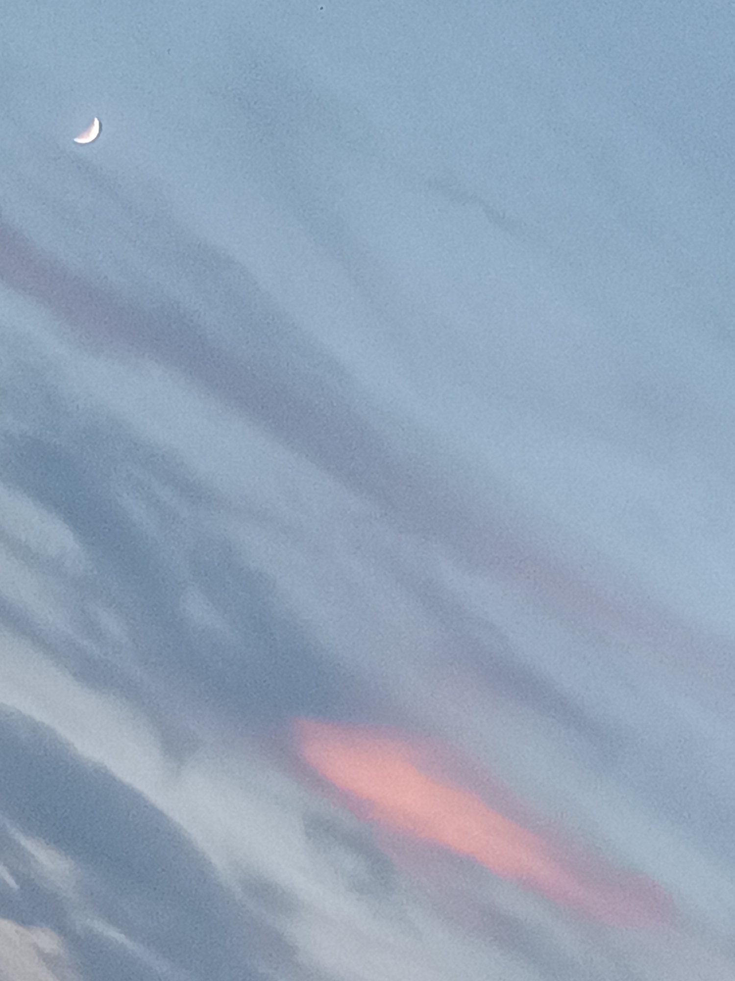 The image shows a sliver of a crescent moon in the upper left quadrant of a pale blue sky. Wispy, diagonal clouds, tinged with a reddish-orange hue in the lower center, stretch across the frame. The scene appears to be an outdoor environment, likely captured during twilight or early evening. The image does not contain any specific landmarks or features that would indicate its location within Viadana, Italy.