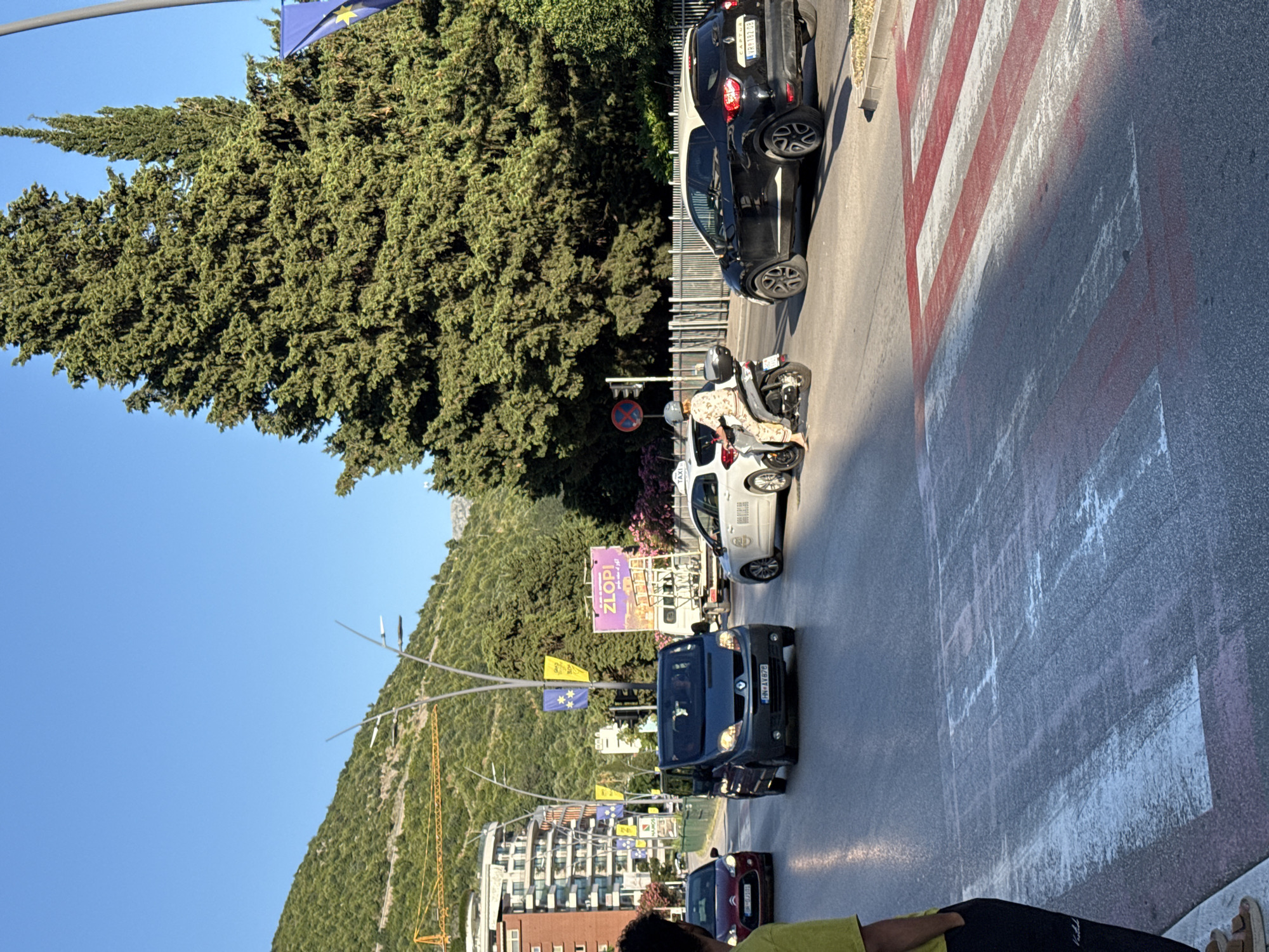 In Budva, Montenegro, on a sunny day, vehicles navigate a street with a pedestrian crossing marked by red and white stripes. A dark blue van, license plate HN-AV678, is in the left lane, followed by a red car. A white taxi, with a rider on a scooter behind it, is in the right lane. A black car is to the right of the taxi. Tall evergreen trees and a distant green mountain form the backdrop. Buildings and streetlights are visible on the left side of the road, and banners with yellow and blue stars hang from poles. The sky is a clear, bright blue.