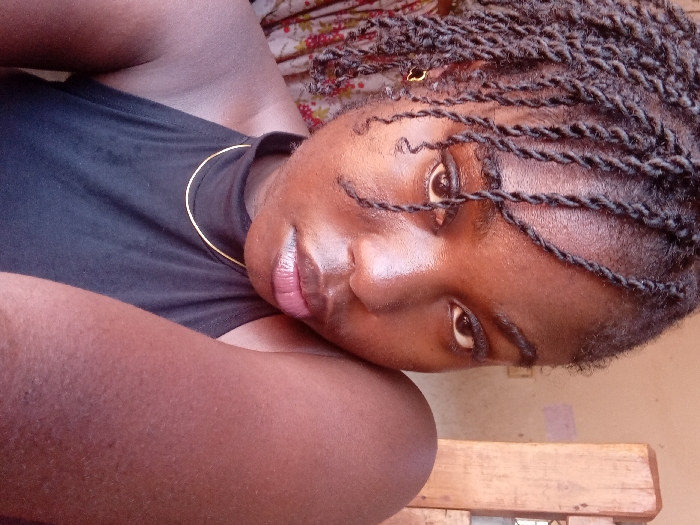 This image is a close-up portrait of a young adult female, taken in an indoor setting in the city of Ejule, Nigeria.
The subject has dark skin and dark, twisted braids, with several strands hanging across her forehead. Her dark eyes are accentuated with eyeliner, and she maintains a direct gaze towards the camera. Her lips are full with a light pinkish hue. She wears a black halter-neck top and is adorned with a thin gold-colored chain necklace. A small, gold-colored earring is visible on her left earlobe. Her right shoulder and upper arm are partially visible in the lower right foreground.
The background consists of two distinct areas. To the left, a patterned fabric, possibly a curtain or textile, with red and white floral or abstract designs is partially visible. To the right, a plain, light brown wall is present, behind a vertical wooden structure, possibly a door frame or furniture element. The scene is illuminated evenly, suggesting natural light.