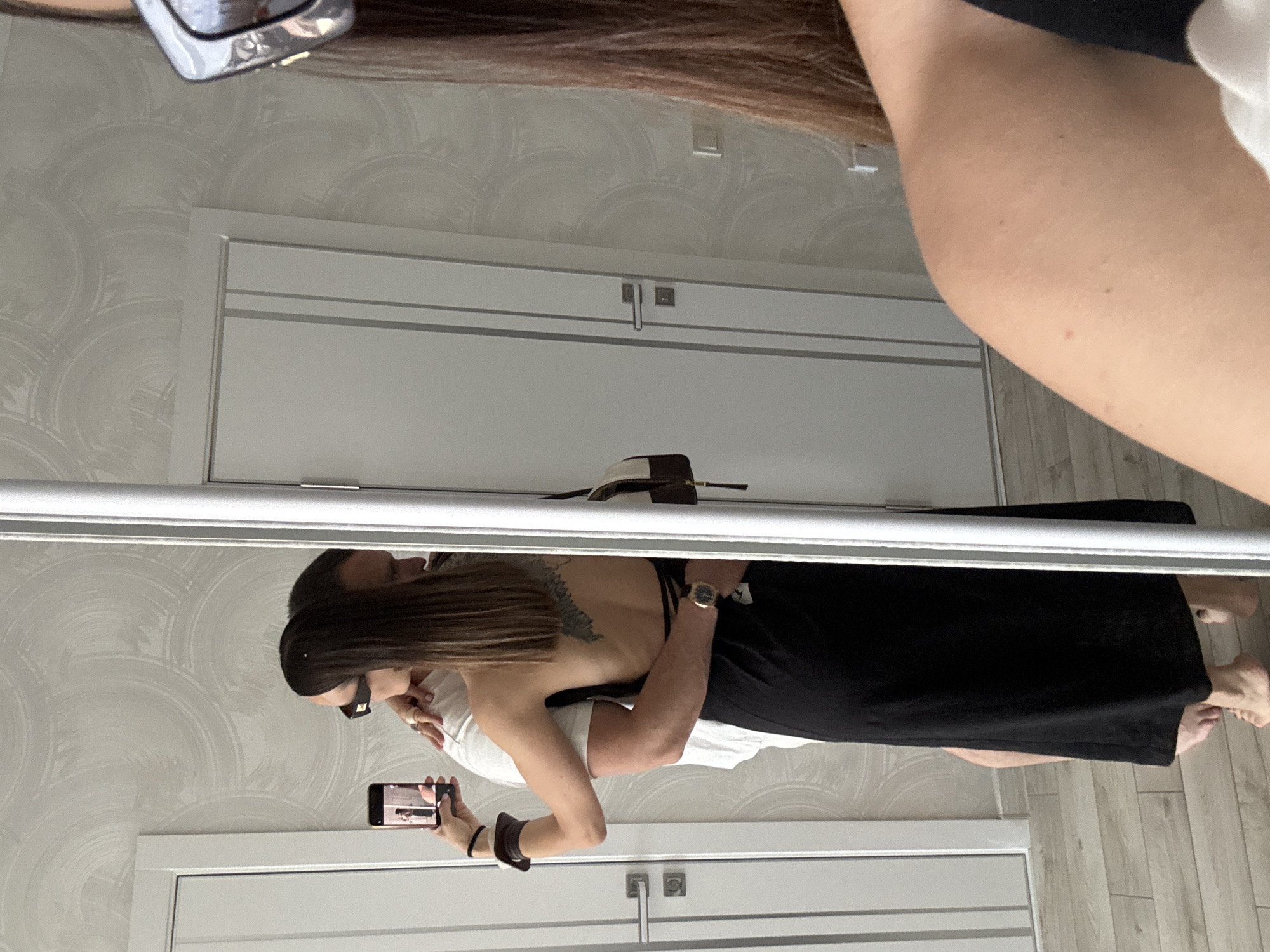 A mirror selfie captures a man and a woman embracing in an indoor setting, identified as a residence in Sochi, Russia.
**People:**
The primary subject is a woman, partially visible in the immediate foreground on the right, and fully visible in reflection within the mirror. She has long, straight brown hair, visible over her right shoulder and upper arm, and is wearing large, square-framed silver sunglasses. In the mirror's reflection, she is seen from the back and side, wearing a black, backless maxi dress with thin straps that crisscross and tie at her lower back. A tattoo, resembling wings or a bird, is visible on her upper back. Her left arm is raised, holding a black smartphone with a gold edge, which is displaying the live camera feed, creating a recursive image of the scene. She wears a dark, thin wristband and a wider, brown and white striped cuff on her left wrist. A gold-colored ring is visible on her right hand (resting on the man's shoulder). Her bare feet are visible on the floor.
A man is embracing the woman from behind, his reflection visible in the mirror. He has short, dark hair and is wearing a white short-sleeved t-shirt. His head is turned, resting near the woman's right shoulder. His right arm is wrapped around her waist, and he wears a gold-colored wristwatch with a dark strap on his right wrist. His bare feet are visible beside the woman's.
**Objects and Environment:**
The scene unfolds within a room with light-colored walls covered in a subtle, repeating circular or swirling pattern wallpaper. The flooring consists of light-toned wooden or laminate planks. Two modern, light-gray or white doors with brushed metal handles and horizontal paneling are visible in the reflection, positioned on the left and right sides of the mirrored couple. A white rectangular wall plate, possibly a light switch or electrical outlet, is visible on the wall between the two doors. A brown and off-white patterned shoulder bag is visible hanging on the right side of the man, likely from his shoulder or suspended from an unseen fixture. The primary reflective surface is a large, full-length mirror with a white or light-colored frame, encompassing most of the central and right portions of the image.
