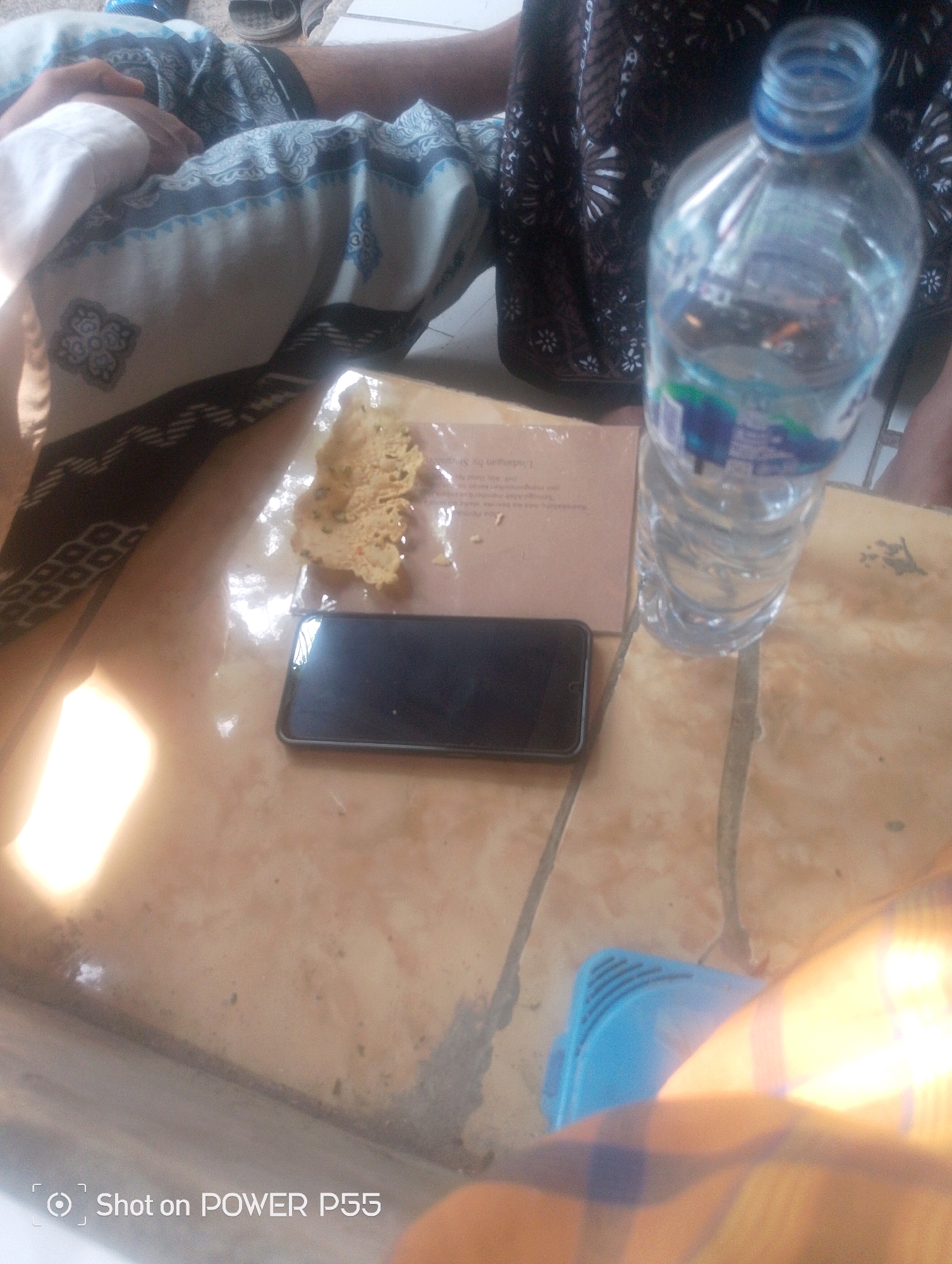 A horizontal, medium shot captures a close-up of a table surface with various objects on it. The surface appears to be a light brown tiled material. In the center of the frame lies a black smartphone. To the left of the phone is a small, irregularly shaped piece of food, possibly a cracker or a crispy snack, resting on a piece of paper. To the right of the phone and food is a clear plastic bottle of water, with a blue cap and label, partially filled. The background is blurred, but suggests people are present, with fabric patterns visible. The lighting appears to be natural, casting a bright glow on the left side of the image, indicating sunlight. The bottom right corner displays a watermark that reads "Shot on POWER P55". The overall scene depicts a casual, possibly outdoor setting.