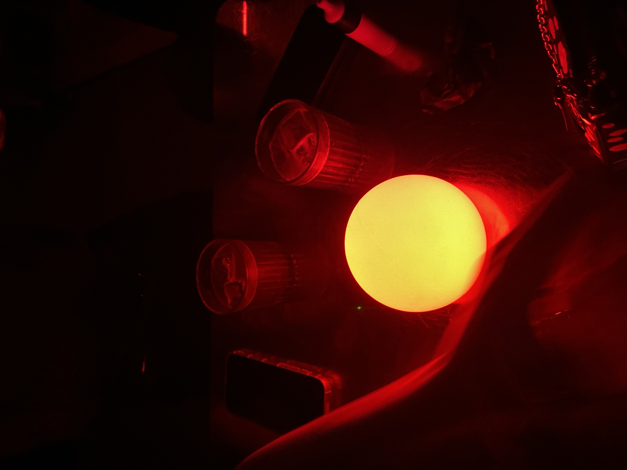 The image shows a dimly lit interior scene with a dominant red hue. A brightly illuminated yellow sphere is the focal point, casting a glow on nearby objects. Two cylindrical glasses, partially filled and appearing red due to the lighting, are positioned to the left of the sphere. A dark, rectangular object, possibly a phone, sits below the top glass. In the upper right corner, a chain and a small, dice-like object are visible. The lower right portion of the image appears to show a person's arm or clothing. The overall environment is dark and indistinct, hindering detailed identification of other objects or people. There is no information to specify that it is in Dubai, United Arab Emirates.