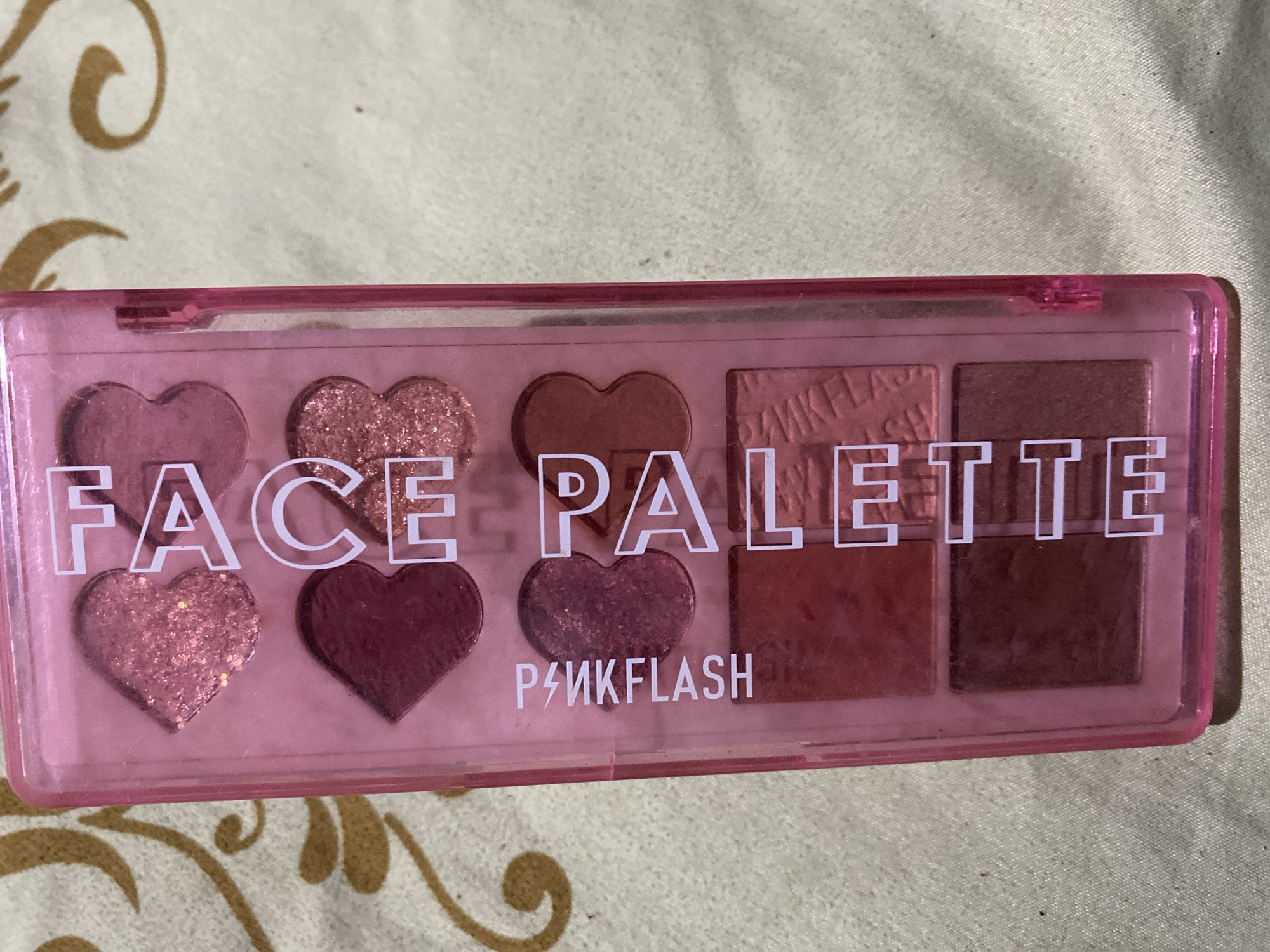 A pink plastic eyeshadow palette is shown from a top-down perspective. The palette is rectangular and has a transparent lid. The lid has the words "FACE PALETTE" printed in white capital letters vertically down the center. To the left of the lettering, "P4MKFLASH" is printed vertically in white capital letters. The palette contains six eyeshadows. The top three eyeshadows are heart-shaped. The top left heart is a sparkly pink. The middle left heart is a deep matte pink. The bottom left heart is a sparkly pink. The bottom right is a rectangle shaped eyeshadow with a textured pattern and a matte finish. The other two eyeshadows are rectangular and have a matte finish in shades of red and brown. The palette is lying on a patterned fabric surface with a light beige background and gold swirls. There are no people or animals present in the image. There is no information about the location within the city of Ta', Indonesia.