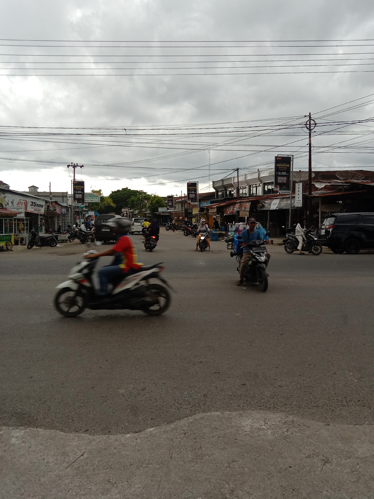 The image depicts a street scene in Lhokseumawe, Indonesia. A blurred motorcycle moves across the foreground. Other motorcycles, both stationary and in motion, are visible in the midground. Buildings line the street, adorned with signage, including advertisements for "SIGNATURE KRETEK" cigarettes. The sky is overcast. Overhead utility lines crisscross the view.