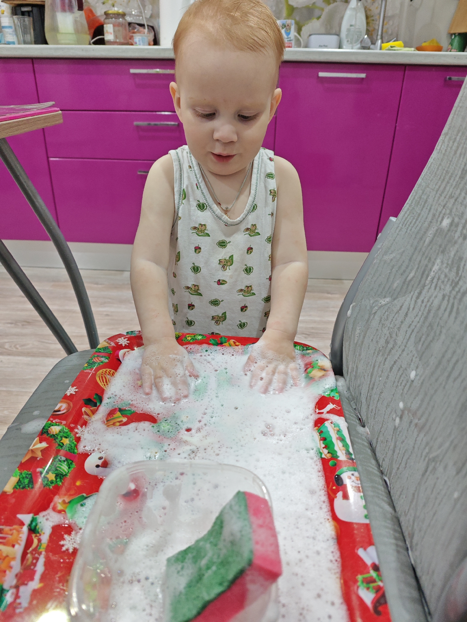 A young child with reddish-blonde hair is the central subject of the image, engrossed in playing with soapy water. The child, likely a toddler, is wearing a white sleeveless top adorned with a pattern of green and brown figures, possibly acorns and small animals or insects. A thin silver chain with a small crucifix pendant is visible around their neck.
The child's hands are submerged in a thick layer of white soap bubbles, which fill a red tray or mat placed on what appears to be a grey highchair or chair. This tray is distinctly decorated with Christmas motifs, including snowflakes, snowmen, Christmas trees, bells, stars, and reindeer, suggesting a festive theme. Inside the bubbly water, a clear plastic container or bowl is partly visible, along with a red and green sponge.
The background shows a domestic setting, likely a kitchen or dining area. Bright pink kitchen cabinets with silver handles dominate the upper left and central background. On the countertop above the cabinets, various kitchen items such as jars and bottles are visible. To the left, a wooden table edge is partially seen, with a pink object resting on it. Part of the grey chair/highchair is visible on the right, lightly splattered with soap. The floor beneath is a light-colored wood or laminate.
The child appears focused and calm, looking down at their hands and the bubbles. The activity seems to be sensory play with water and soap, possibly mimicking washing, in a home environment in Markovskii, Russia. There is no visible text in the image.