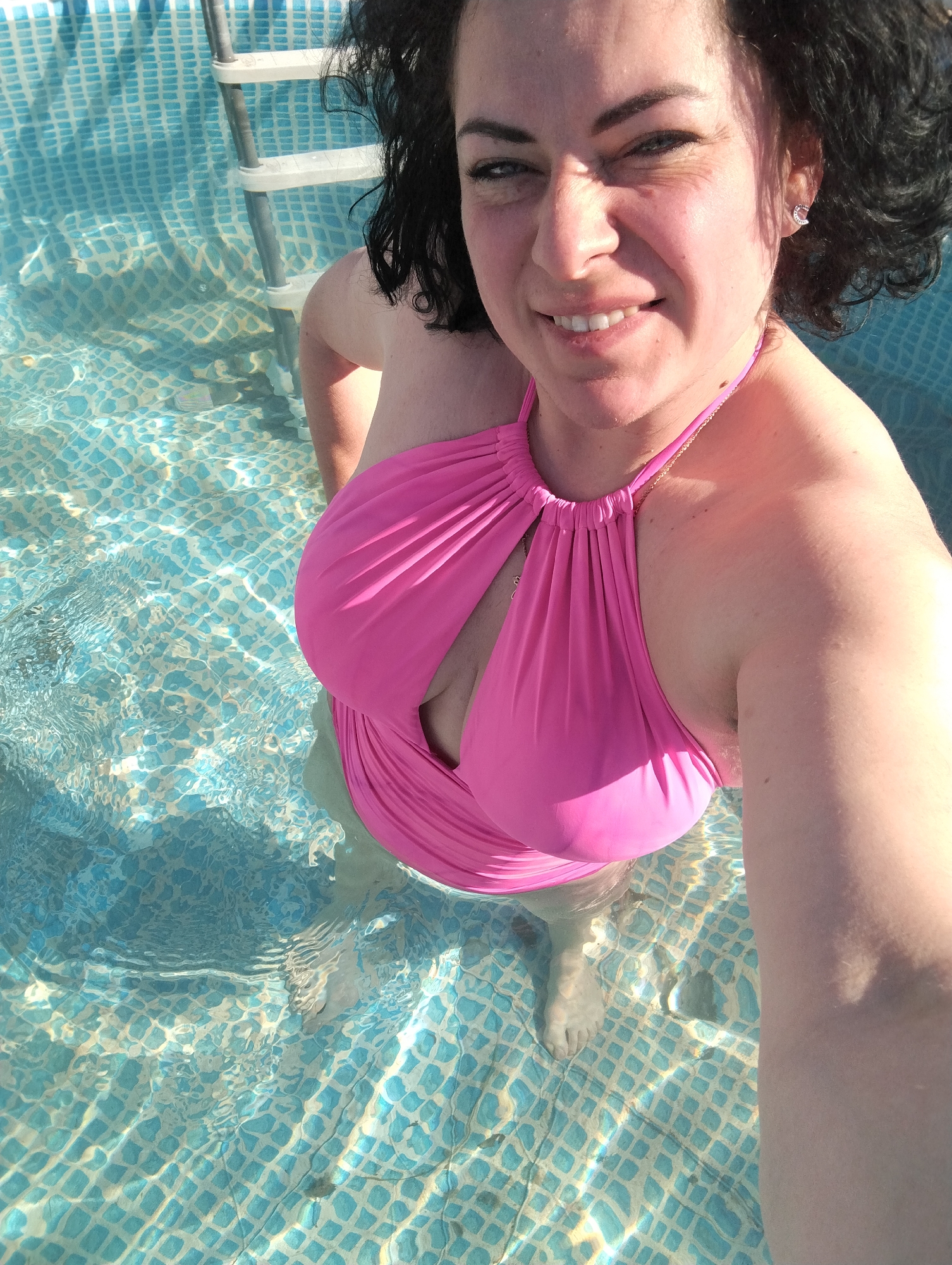 A woman is standing in a swimming pool with light blue tiled walls and floor. She is wearing a pink halter-neck swimsuit. The water level reaches her mid-thigh. A metal ladder is visible against the pool wall. The woman is looking directly at the camera with a slight smile. Her hair is dark and curly. The location is not Grishanki, Russia.
