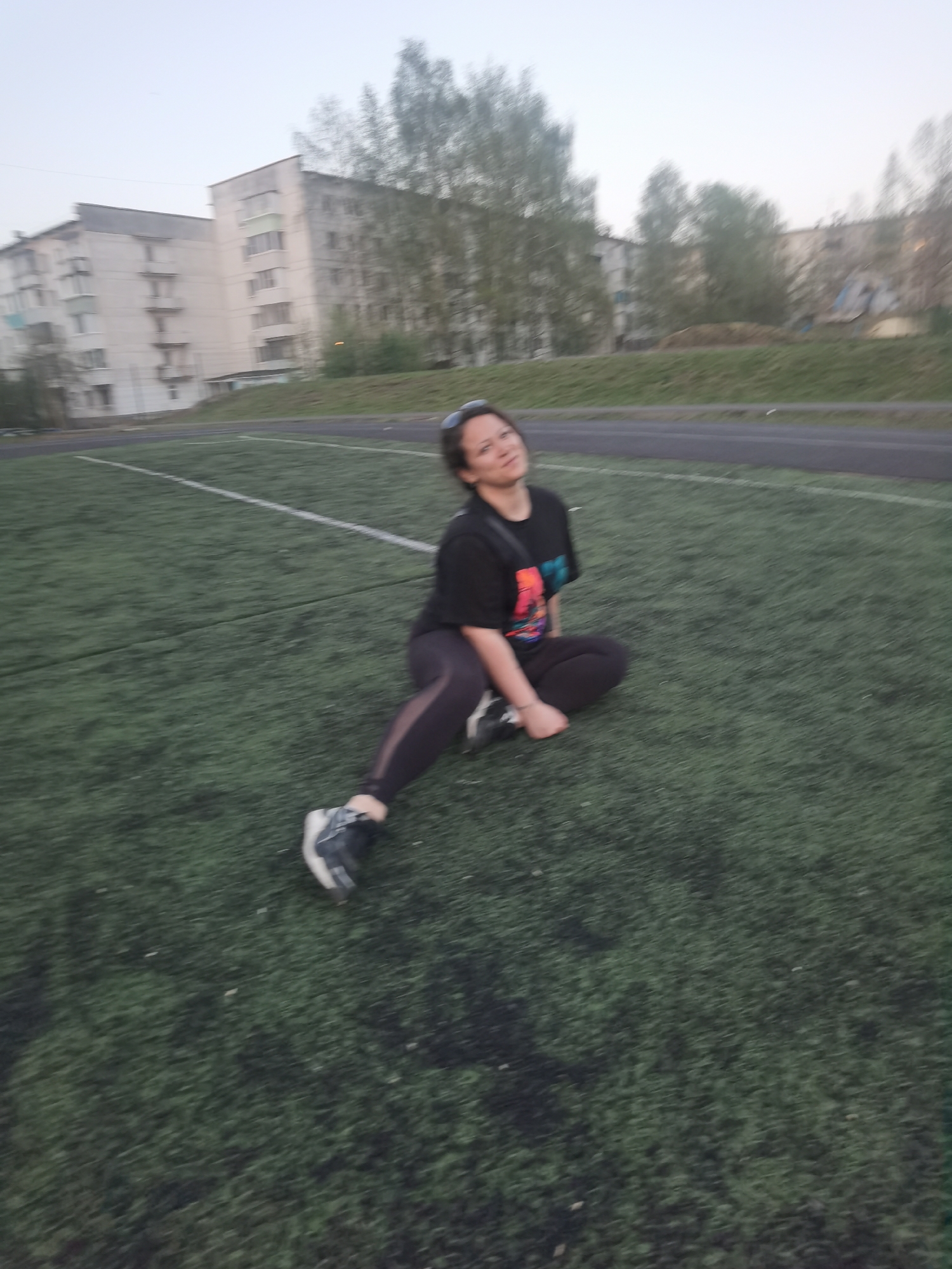 A woman is sitting on an artificial turf field in Chaikovsky, Russia. The field is a green color with white lines, suggesting it is a sports field. The woman is wearing a black t-shirt, dark leggings, and sneakers. She is sitting in a cross-legged position with one leg extended. In the background, there are residential apartment buildings and trees. The weather appears to be overcast.