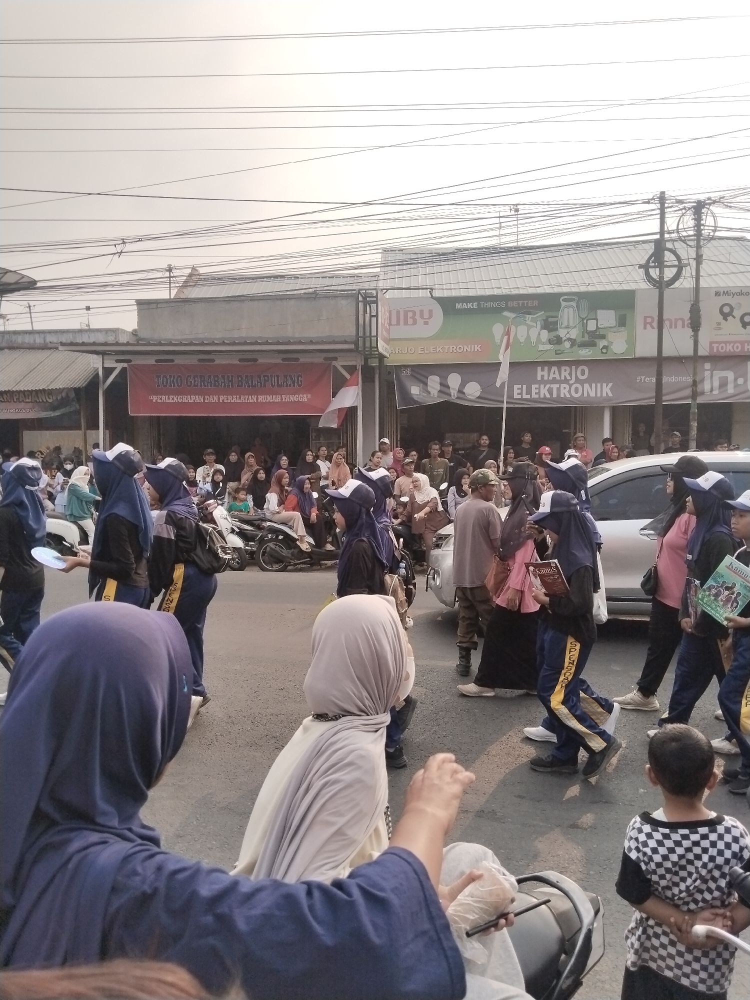 In Balapulang Kulon, Indonesia, a procession is underway on a street lined with shops. A group of individuals, mostly young people wearing blue and dark uniforms with headscarves, are walking in a column. One person in the foreground is wearing a purple headscarf and dark clothing, with their arms outstretched. Another person in the middle ground wears a beige headscarf and light-colored clothing. A young boy in a checkered shirt is standing with his hands behind his back, facing away from the camera. Several motorcycles are parked along the side of the street, and a silver car is visible. A red banner hangs across a building that reads "TOKO GERABAH BALAPULANG" and "PERLENGKAPAN DAN PERALATAN RUMAH TANGGA". Another banner above it displays "HARJO ELEKTRONIK" and "MAKE THINGS BETTER" with images of electronic appliances. The sky is overcast, and overhead power lines are prominent. The scene appears to be a daytime event, possibly a parade or a school event.