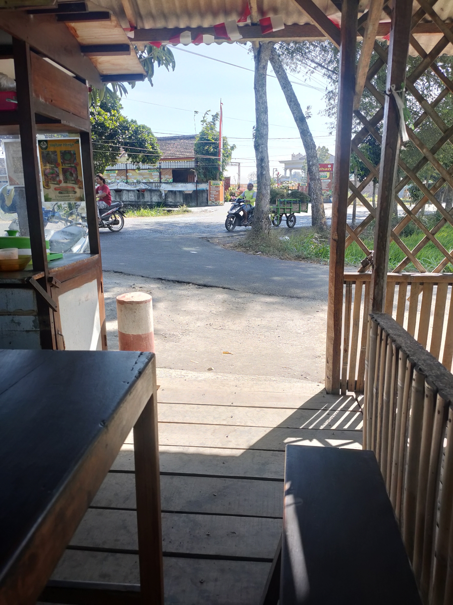 The image captures a view from a covered wooden structure, possibly a roadside stall or eatery, looking out onto a street in Kanigoro, Indonesia. In the foreground, a dark wooden table and bench are visible, with sunlight casting striped shadows on the wooden deck floor.
Outside the structure, a road is present with two individuals on motorcycles. The first, a person wearing a red shirt, is riding a motorcycle from left to right. Further down the road, another person in a lime green shirt is operating a motorcycle with a trailer attached, carrying a load of green vegetation. The street is bordered by a sidewalk and then grass and trees. In the background, several buildings with tiled roofs are visible, along with more trees and vegetation. The sky is clear and blue. A red and white striped marker is positioned near the edge of the road. The overall impression is of a sunny day in a moderately developed area.