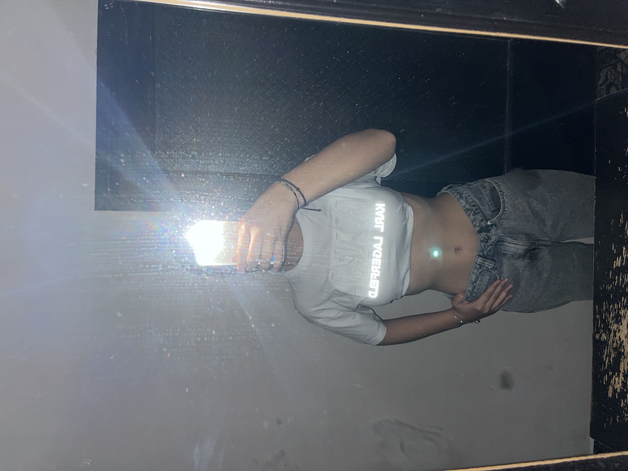 The image shows a person taking a selfie in a mirror. The person is wearing a white Karl Lagerfeld crop top and grey jeans. The background is a wall with a black tiled pattern. The location is Unknown, Unknown. The person is holding a phone in front of their face, blocking their face from view. The reflection in the mirror is the primary subject of the image.