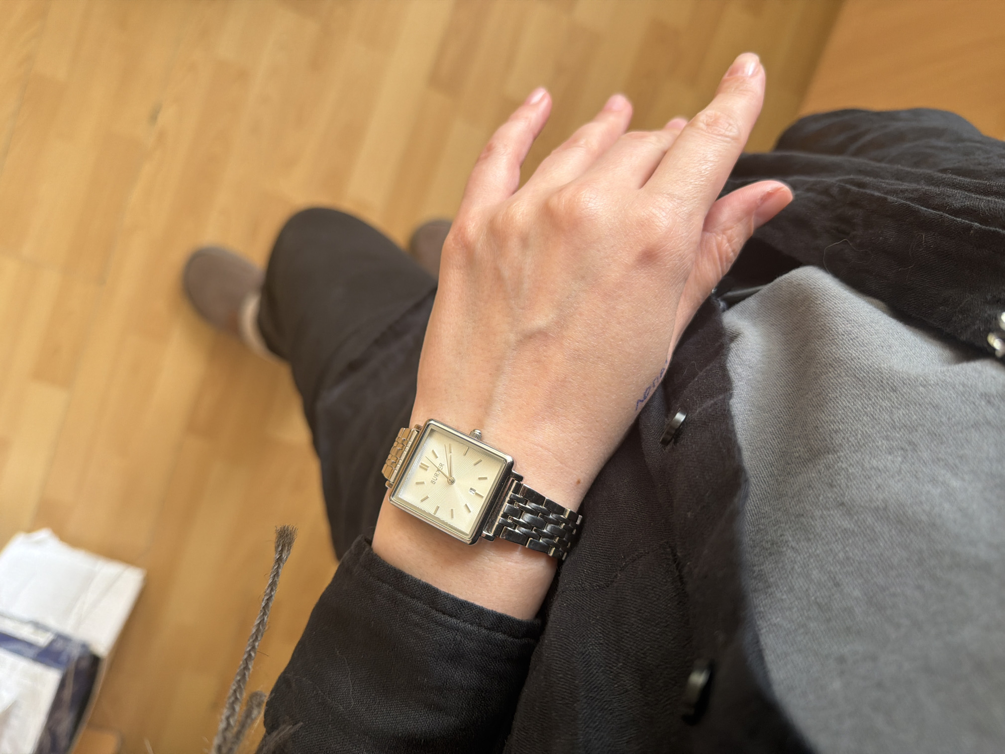An indoor scene in Chaikovsky, Russia, depicts a person's upper legs, left arm, and hand from a first-person, downward perspective. The individual is seated, wearing dark-colored trousers and a long-sleeved dark shirt or jacket, with a lighter grey fabric visible on the lap. On the left wrist is a rectangular, silver-toned wristwatch featuring a metal link bracelet, a light-colored face with radial grooving, numerical hour markers, and a date window at the 6 o'clock position; the brand "BURBER" is partially visible. The person's fair-skinned left hand rests on the lap. In the blurred background, a light brown wooden floor is visible. To the upper left, a white package or item rests on the floor. A grey braided cord or drawstring, possibly part of the clothing, hangs into the frame from the upper left. A grey slipper with a light-colored trim is visible on the person's right foot in the lower right background.