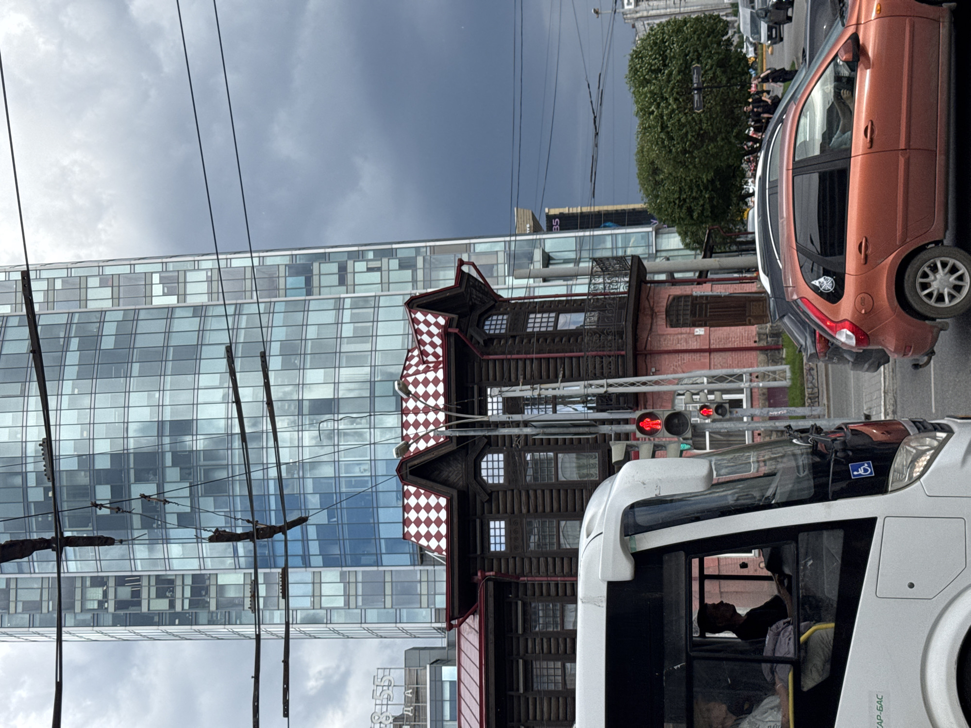 The image depicts an urban scene in Yekaterinburg, Russia, characterized by a contrast between modern and historic architecture. A tall, glass-fronted skyscraper dominates the background, reflecting the overcast sky. In the mid-ground, a traditional wooden building with a red and white checkered roof stands out. A white bus is partially visible in the foreground, with its driver in view. Several cars are present on the road, indicating traffic. The overall atmosphere suggests a cloudy day in a bustling city.