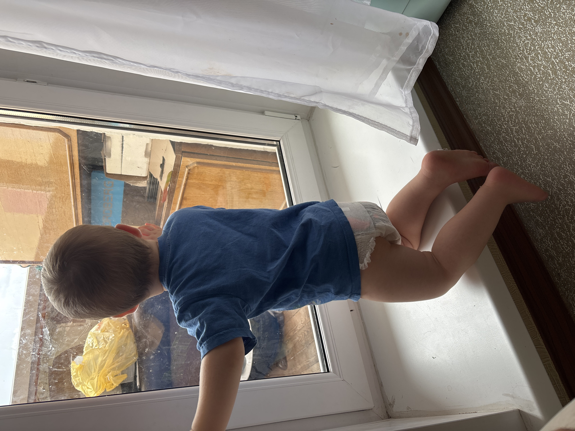 A young child, wearing a blue t-shirt and a diaper, is in a crawling position on a white windowsill. The child is facing away from the camera, looking out through a glass door or window. The view outside appears to be a cluttered area with wooden structures and a yellow plastic bag visible. The scene is indoors, with a white curtain or sheet hanging above the window. The floor beside the child is covered in a textured, speckled wallpaper and a dark wood trim. The lighting suggests it is daytime. The image appears to be taken in Naberezhnye Chelny, Russia.