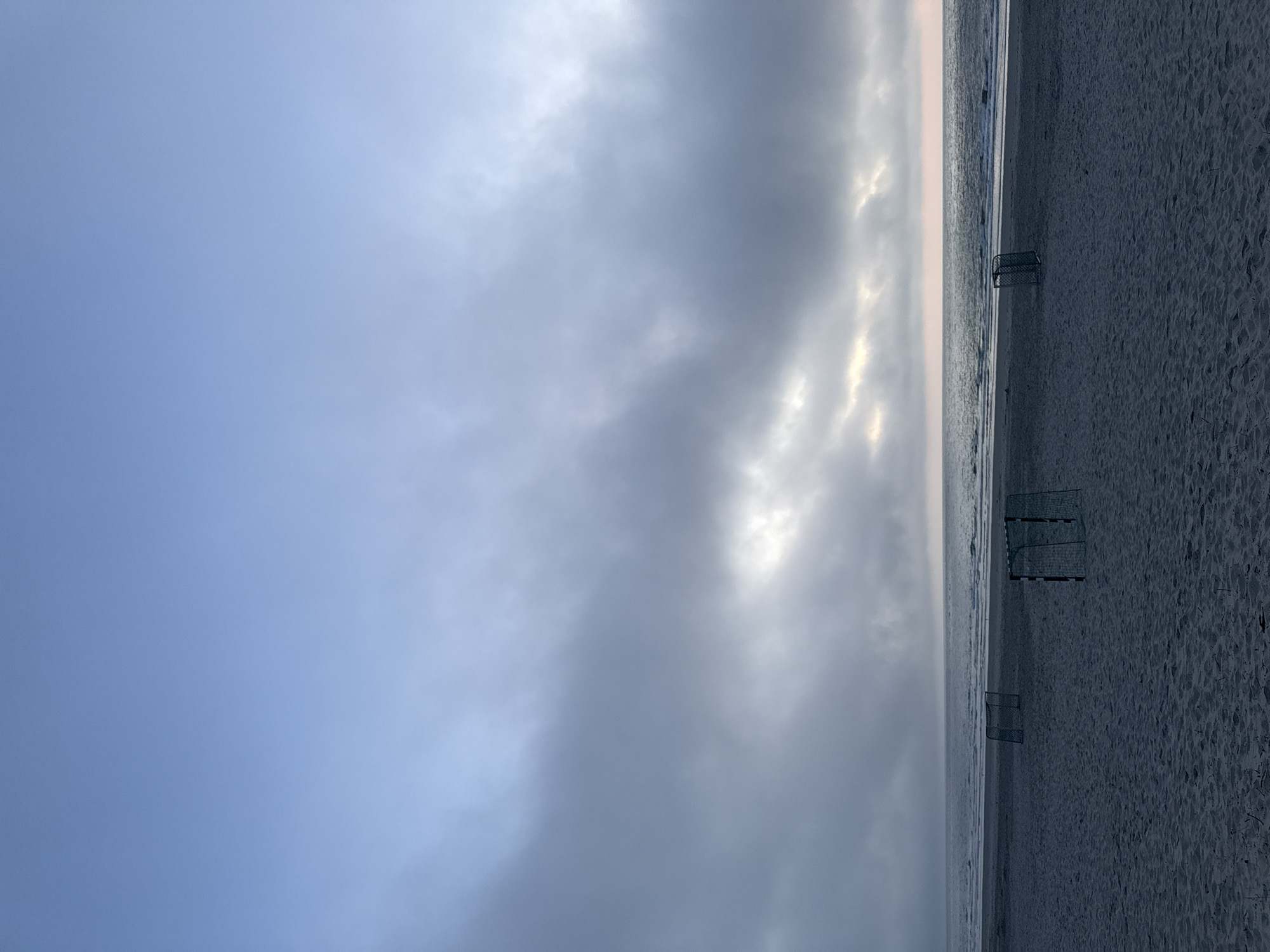 The image depicts a sandy beach on the island of Langeoog, Germany, under a cloudy sky. In the foreground, three small, portable goal frames, likely for beach sports, are visible on the sand. The sea stretches out towards the horizon, with gentle waves lapping at the shore. The sky is a gradient of blues and grays, with patches of brighter light suggesting the sun attempting to break through the clouds. The overall atmosphere is calm and somewhat overcast.