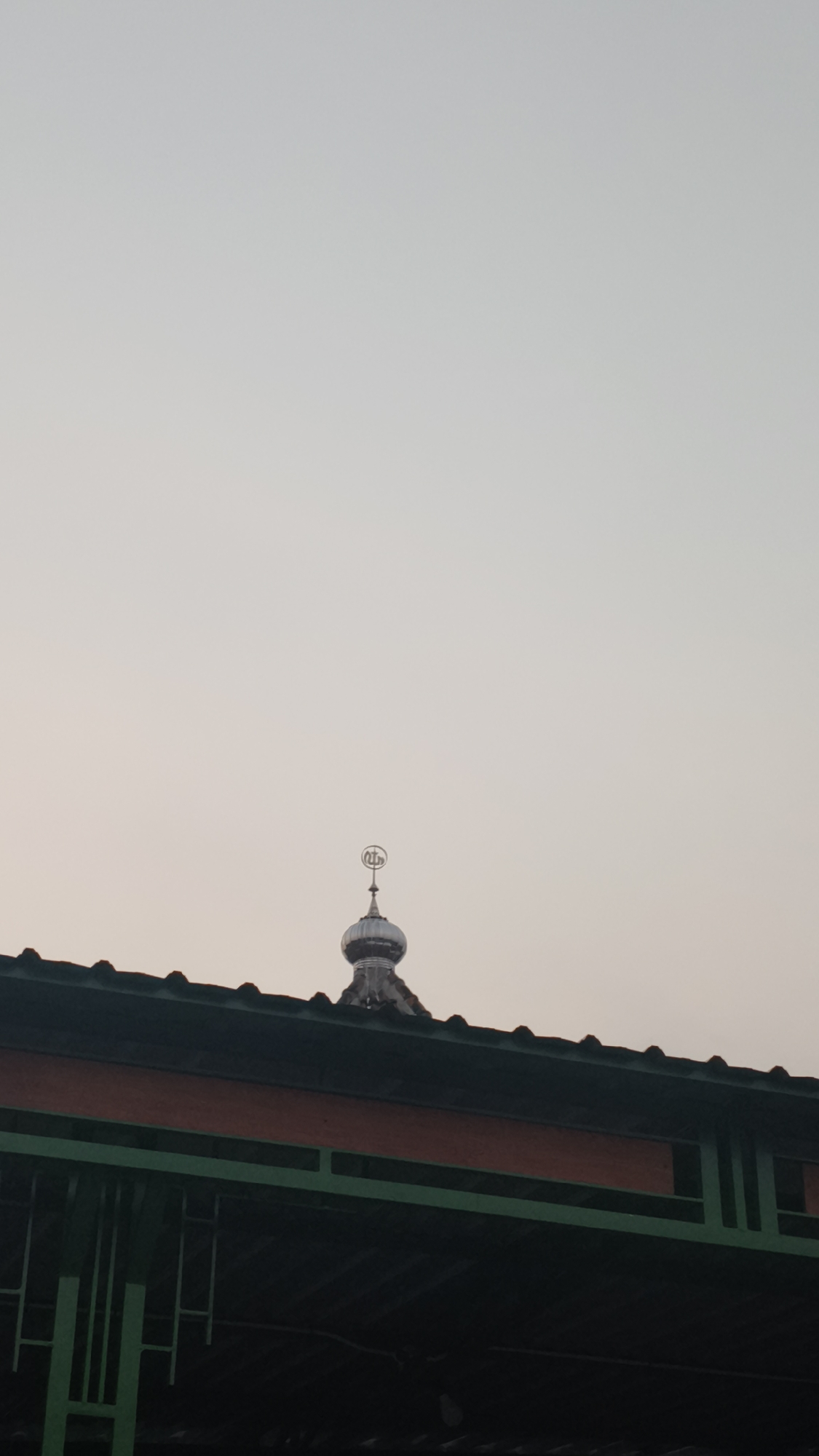 A close-up, low-angle shot captures the top of a building, likely a mosque, in Kerep Kidul, Indonesia, against a pale, overcast sky. The building's dark, sloped roof is visible along the bottom edge of the frame, bordered by a bright orange band and green architectural elements. Perched atop the roof is a gleaming, silver dome topped with a crescent moon and star finial, symbolizing Islam. The overall impression is one of quiet solemnity and religious significance.