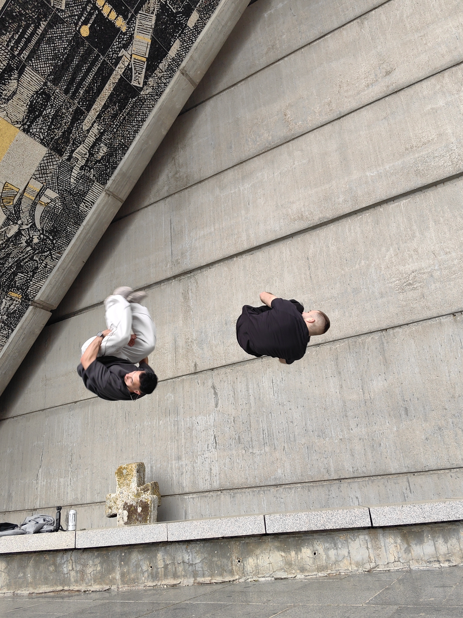 The image displays two men performing acrobatic maneuvers, likely backflips or tuck jumps, against a large concrete structure. The photo is taken from a dynamic, tilted perspective, making the expansive concrete wall on the right appear vertical.
The man on the left is mid-air, wearing a black t-shirt and white athletic pants, with his body tightly tucked as he rotates. His face is visible, looking downwards. Further to the right, a second man in a black t-shirt and dark pants is also airborne in a similar tucked position.
The background consists primarily of a textured concrete wall with horizontal panel lines. To the upper left, an angled concrete beam connects to a section of wall adorned with an intricate mosaic or mural. This mural features abstract or geometric patterns in black, white, and hints of gold or yellow.
At the bottom of the frame, a stone ledge runs horizontally, topped with granite-like slabs. On this ledge, a weathered, moss-covered stone cross-shaped object is visible. Alongside it are a gray backpack or bag, a white can with the word "MONSTER" clearly visible, and a small, portable camera or gimbal. Below the ledge, there is a lower concrete wall with cracks and a darker ground surface.
The lighting indicates daytime conditions, possibly overcast, as there are no strong shadows. The setting appears to be an urban or public architectural space, characterized by concrete and stone elements, possibly an underpass or a public plaza with distinctive artistic features. The scene captures a moment of intense athletic activity in an unconventional architectural environment.