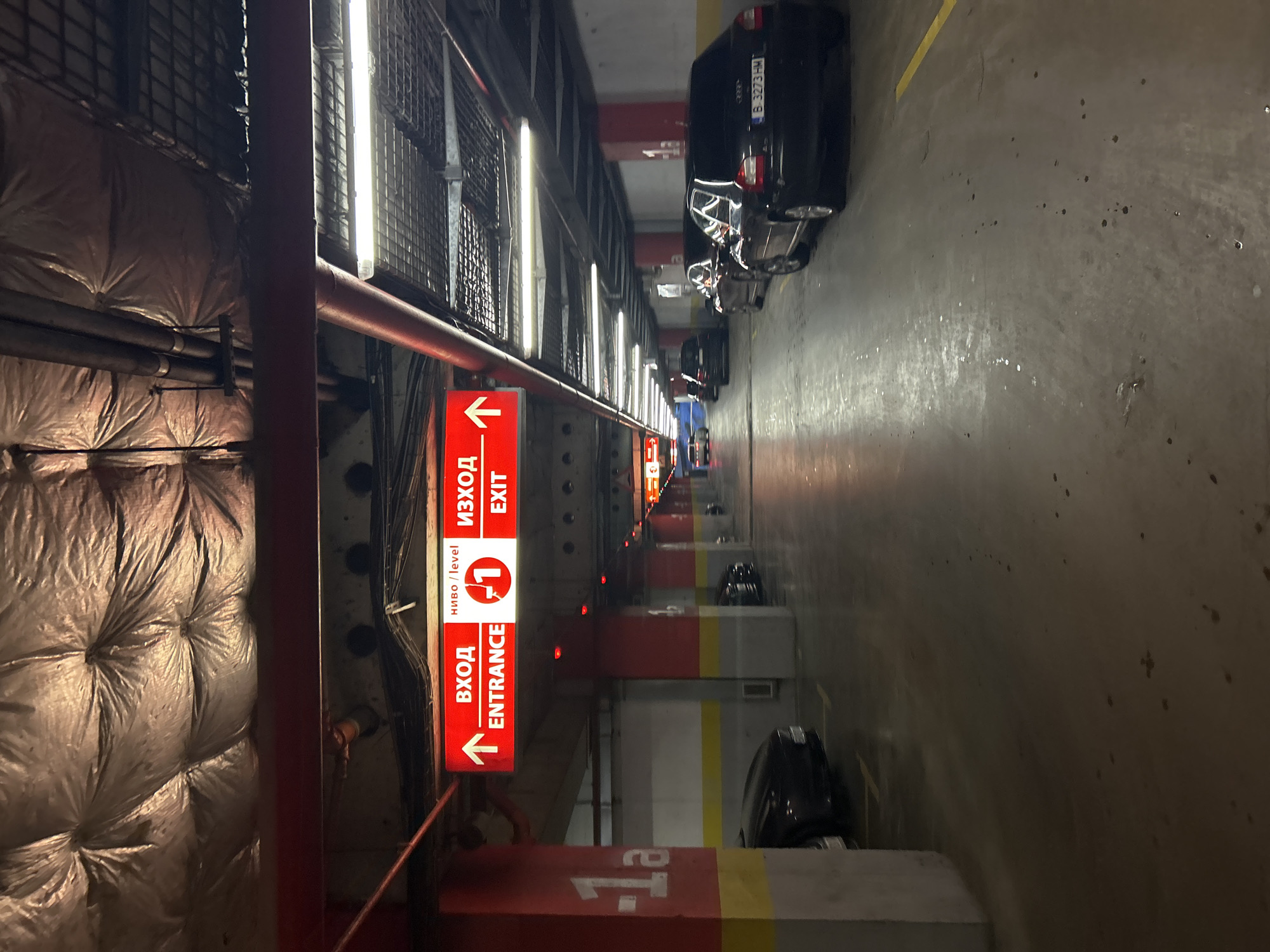 The image depicts an indoor parking garage in Varna, Bulgaria. A red and white sign labeled "Вход Entrance, Ниво/Level 1, Изход Exit" indicates the entrance and exit to the garage's first level. Cars are parked along the parking spaces, which are defined by red and yellow painted markers and white concrete curbs. The floor is concrete with dark splotches. Overhead, there are rows of fluorescent lights, pipes, and a metal grid. One side of the garage features insulated wrapping on the walls. The scene shows a typical, functional parking area.