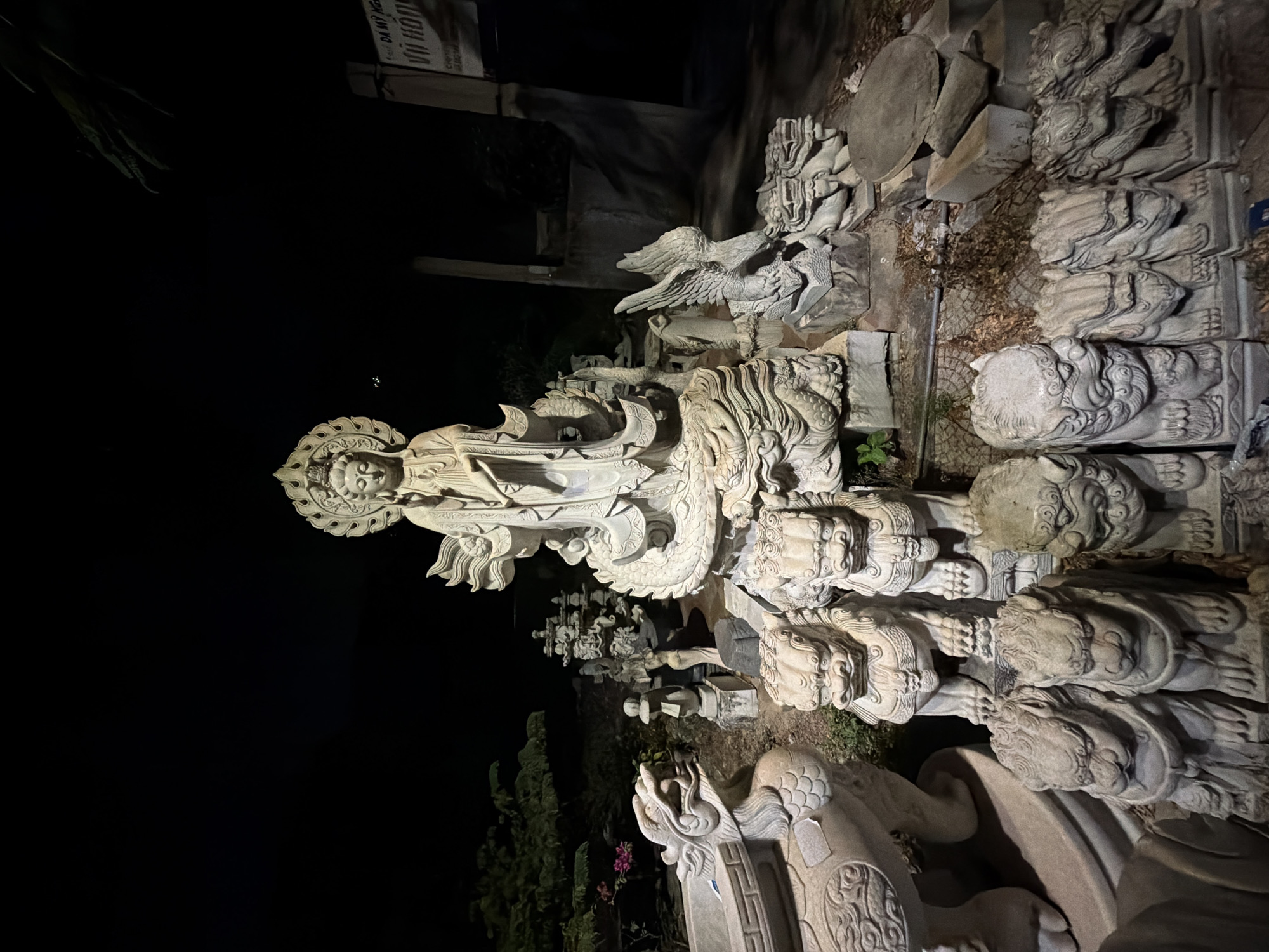 This is a night shot depicting a collection of stone sculptures, primarily Buddhist in nature, likely located in Đà Nẵng, Vietnam. The most prominent sculpture is a statue of a deity with a halo, standing atop a serpent or dragon motif. There are numerous other carved stone elements, including lion-like figures, and winged mythical creatures. The statues appear to be in a stone yard, potentially a workshop or storage area. There are trees visible in the background. The lighting is uneven.