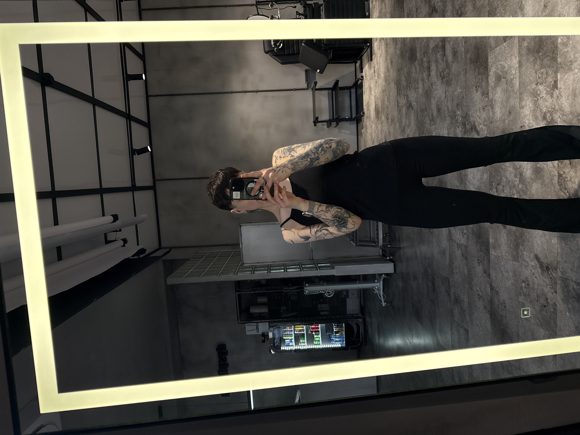 A full-body mirror selfie shows a person with short dark hair, tattooed arms, and dressed in black clothing, holding a smartphone to take the picture. The reflection reveals the interior of what appears to be a commercial establishment or salon, with overhead lighting fixtures and dark floors. In the background, there is a vending machine stocked with colorful items, as well as various pieces of furniture possibly related to the salon. Without further information, it is not possible to confirm whether this photo was taken in Moscow, Russia.