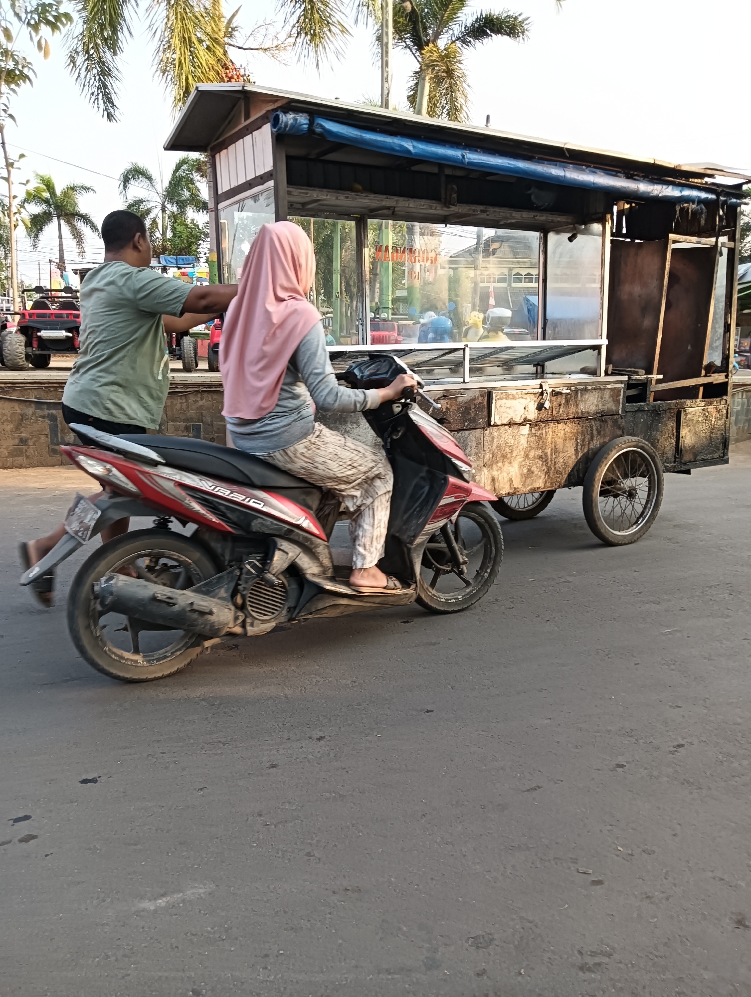 In Tayu, Indonesia, a man in a green t-shirt pushes a cart attached to a motorcycle. A woman wearing a pink hijab is riding the motorcycle, which is pulling the cart. The cart appears to be a food stall with a metal roof and glass display cases. The scene is set on a paved road with palm trees in the background. Red ATVs are visible behind the food cart.