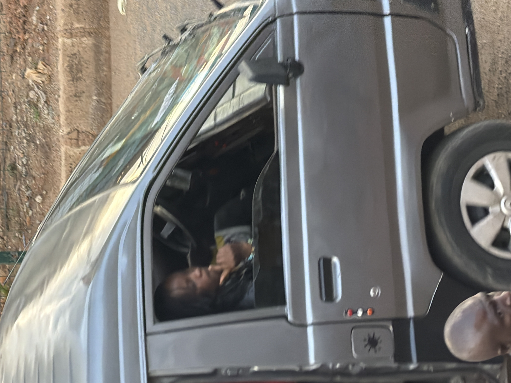 A medium shot captures a side view of a gray minivan, with a person sitting in the driver's seat. The individual, with dark hair, appears to be looking down and resting their chin on their hand. The minivan's interior is partially visible, including the steering wheel. Outside the vehicle, a dirt road and a textured, reddish-brown curb are visible. In the foreground, a bald person's head is cropped into the frame. The scene is set in Bukuru, Nigeria.