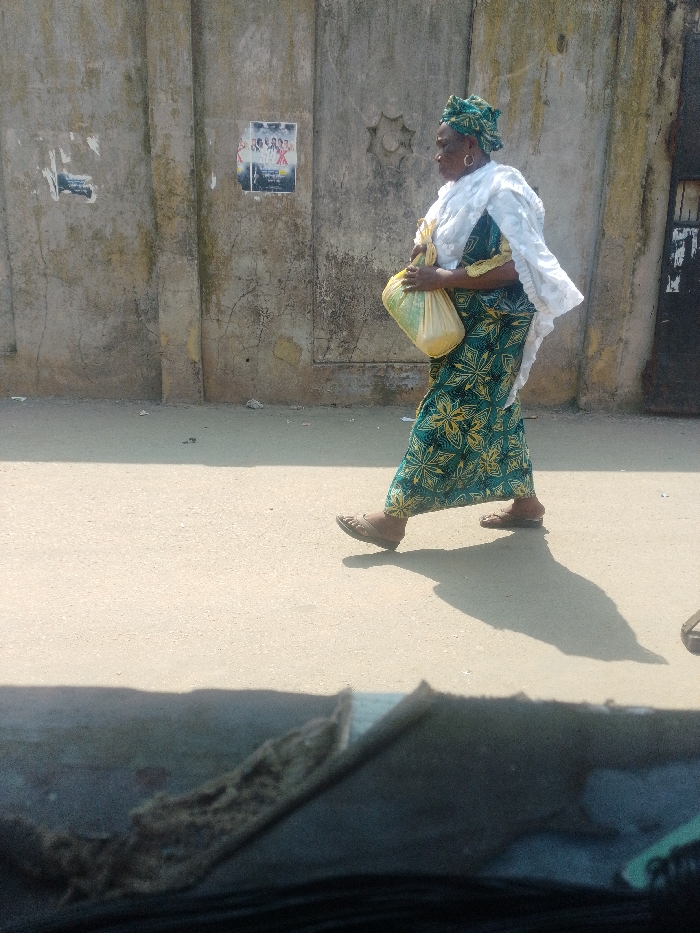 An outdoor scene in the city of Agani, Nigeria, depicts an adult female walking rightward on a light-colored, unpaved road during daylight hours under direct sunlight. The individual, dressed in a long, patterned teal and yellow garment, a white shawl, and a matching headwrap, carries a translucent yellow plastic bag in her left hand. She wears open-toed sandals. Her elongated shadow is cast on the ground to her right. The background features a weathered, light grey concrete wall with visible cracks and discoloration. Affixed to this wall are at least two posters, one prominently displaying images of multiple people in what appears to be an advertisement. A star-shaped decorative emblem is visible on the wall to the right of the main poster. The extreme foreground shows a blurred, dark object, possibly part of a vehicle's interior.