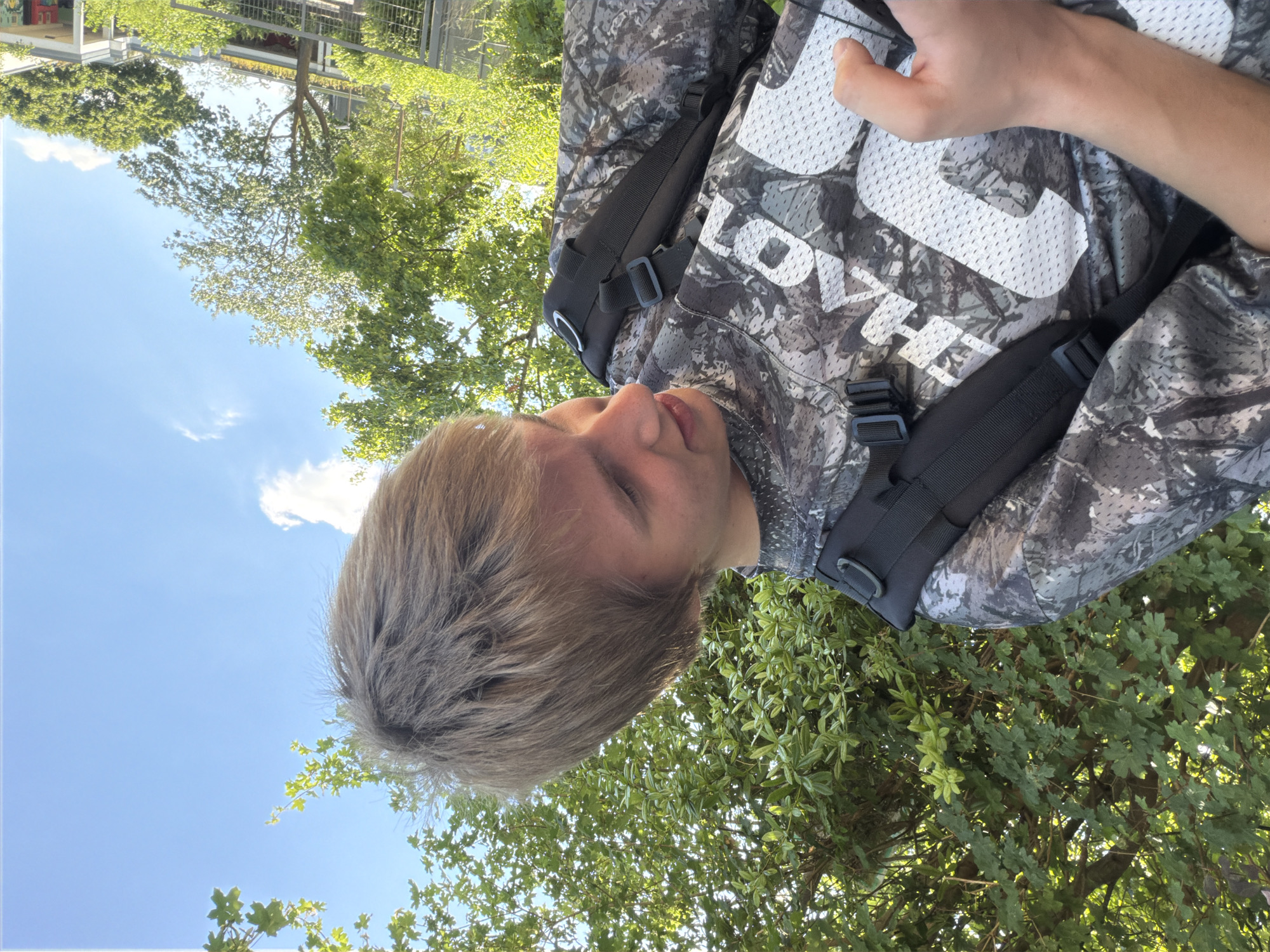 The image shows a person with blonde hair wearing a camouflage-patterned shirt and a black backpack. The person is looking upwards, and their face is partially visible. The background consists of green trees and a bright blue sky with a few white clouds, suggesting it is daytime and the weather is fair. The perspective of the photo is from below, looking up at the person and the sky. There is no discernible activity taking place beyond the person being present in the scene. The image appears to be taken outdoors in a natural environment with trees. The text visible on the person's shirt is white and appears to be letters, possibly part of a brand name or logo, but it is not entirely legible due to the camouflage pattern and the angle. No specific location cues beyond the presence of trees and sky are visible, although the prompt provides the location context of Dorsten, Germany. The lighting suggests it is likely daytime, possibly midday given the brightness of the sky.
