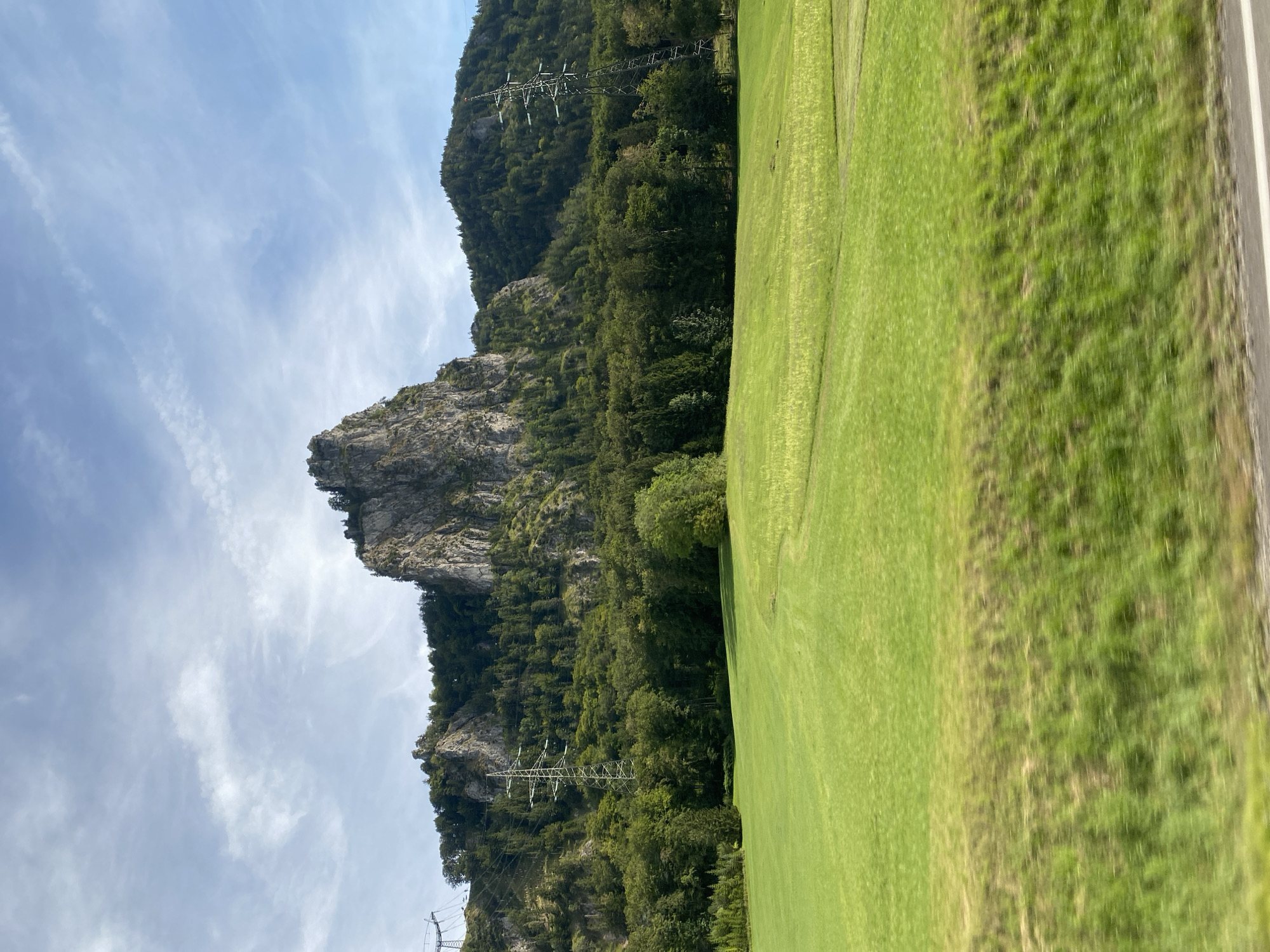 A wide, angled outdoor shot captures a mountainous landscape in Heuberg, Austria. In the foreground, a verdant, mown grass field slopes gently towards the viewer. The field exhibits slight undulations and subtle texture variations, with some areas appearing lighter green or showing remnants of cut grass.

Dominating the midground and background is a lush, dense forest composed of various green trees, creating a rich tapestry of foliage. Interspersed within the forest and on the slopes of the mountains are rocky outcrops and sheer cliff faces, characteristic of the alpine terrain. The central peak is particularly striking, with jagged, weathered rock formations.

Several tall, metallic electrical pylons are visible, some extending high into the sky, suggesting the presence of power lines that traverse the landscape. The sky above is a pale blue, adorned with scattered white clouds that are wispy and elongated, indicating a partly cloudy day. The overall impression is one of a natural, rugged environment with elements of human infrastructure.
