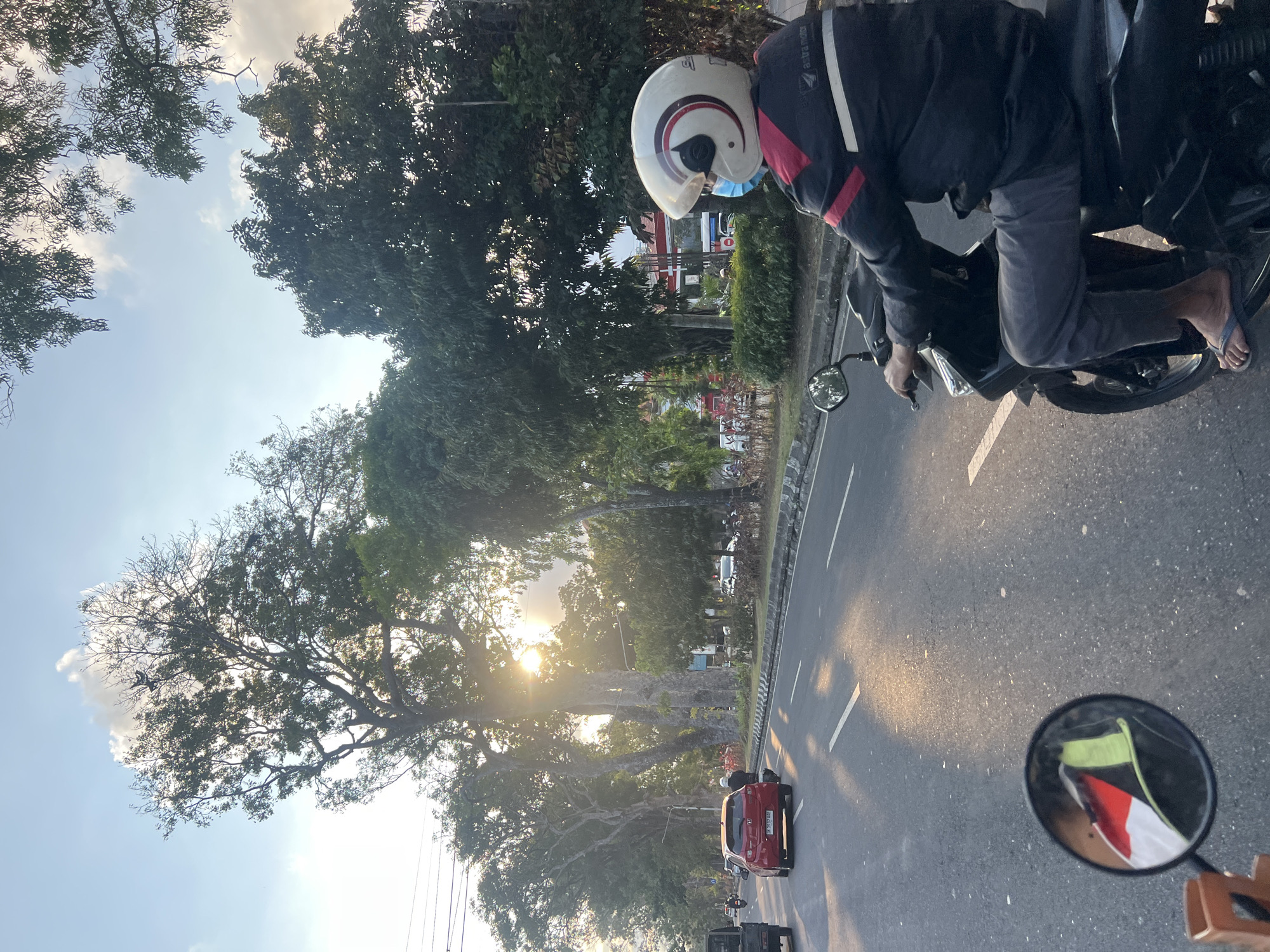 This image captures a street scene in Denpasar, Indonesia, bathed in the warm glow of the late afternoon sun. The perspective is from a moving motorcycle, with the rider in the foreground wearing a white and red helmet, a blue mask, and a black and red jacket. The rider is operating the motorcycle with their right hand on the handlebar and their left hand is not visible. Their legs are extended forward, and they are wearing grey pants and flip-flops.
In the rearview mirror of the motorcycle, a reflection shows a small Indonesian flag with red and white bands. The road ahead is paved and marked with white lines. Several vehicles are visible on the road, including a red car with Indonesian license plates (DK 3937 AAE) in the middle lane, a black truck on the left side, and other vehicles further down the road.
Lining the street are numerous large trees with lush green foliage, casting long shadows on the asphalt. The sun is visible through the branches of some of the trees, creating a dappled light effect and a lens flare. In the background, there are buildings and more trees, suggesting a well-developed urban area within the city. The sky is a mix of blue and light clouds, indicating fair weather. The overall impression is of a typical day on the road in Denpasar.