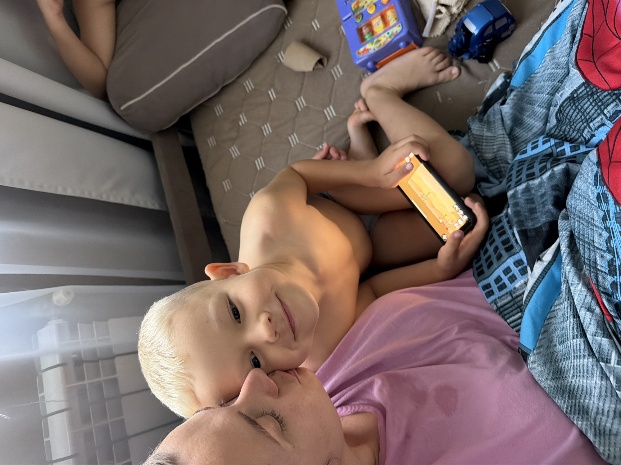 The image captures an intimate, high-angle view of an adult and a young child lying closely together on what appears to be a bed or large sofa, likely indoors in Sochi, Russia. The setting suggests a casual, relaxed home environment.
The adult, with fair skin and visible eyelashes, has their eyes closed, implying rest or sleep. They are wearing a light purple shirt, which shows a small damp spot near the bottom. The child, with blonde hair and fair skin, is lying on their side, looking directly at the camera with a joyful smile. Their head is gently resting near the adult's face, indicating a close bond.
The child holds a black smartphone horizontally, displaying a colorful, cartoon-style game or video on its screen, featuring a light-colored landscape. The child's bare arms and torso are visible, along with light-colored underwear.
Surrounding the subjects are various items: a blue, grey, and red patterned blanket or duvet, possibly Spiderman-themed, is draped on the right. Towards the top, a blue toy car and a purple children's toy resembling a vending machine with colorful compartments are visible. Grey and beige patterned pillows are also scattered on the surface. A crumpled piece of light-colored material lies near one of the pillows. The lighting suggests daytime, possibly with natural light entering from a window on the left side of the frame. The overall mood is one of peace, affection, and contentment. No discernible text is visible within the image.