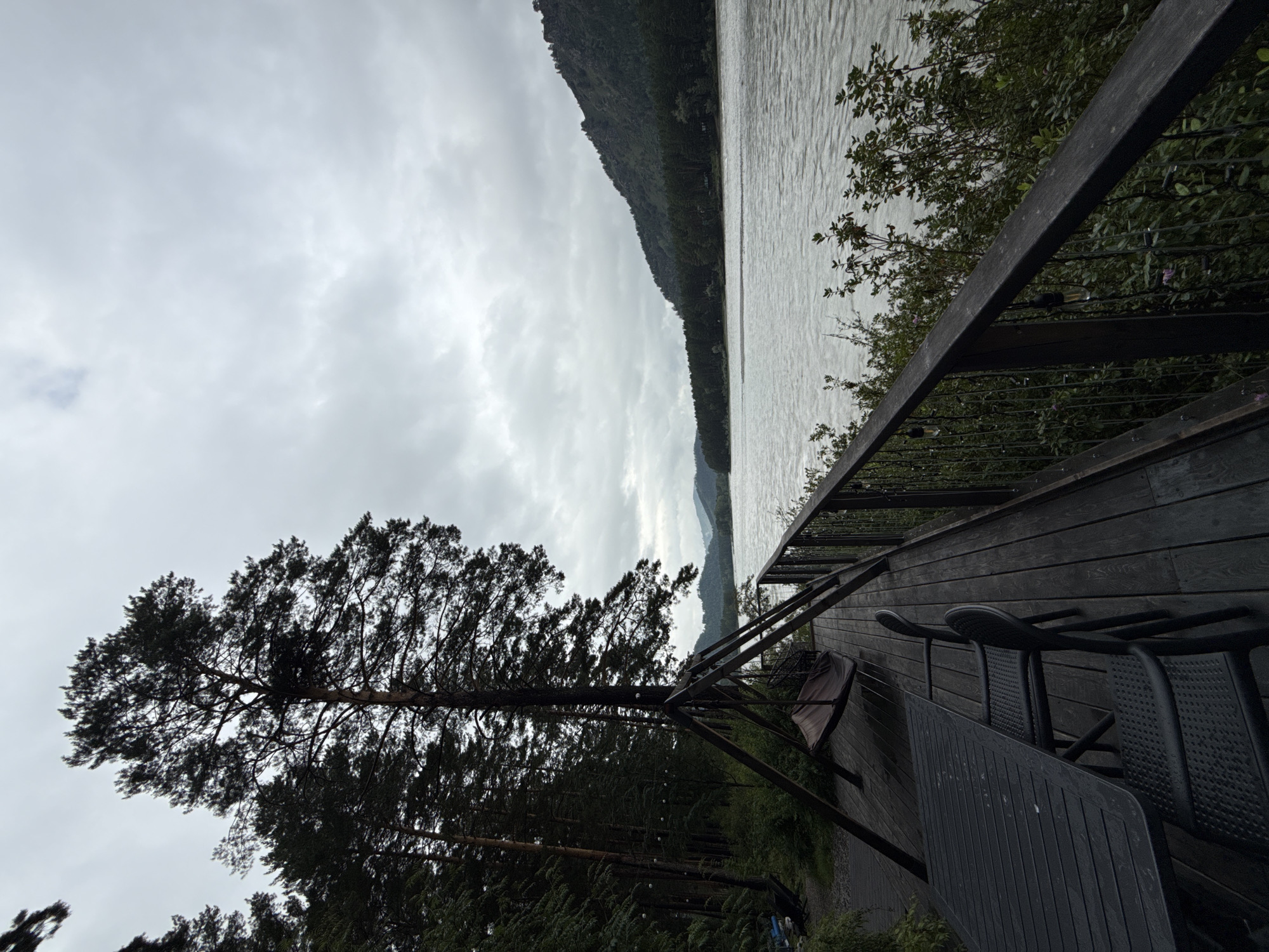 A wooden deck overlooks a wide, fast-flowing river. On the deck, a black table and chairs are set up, suggesting an outdoor dining area. Further along the deck, a swing hangs from a metal frame, positioned with a view of the river and the forested mountains beyond. The sky is overcast, casting a muted light over the scene. The surrounding environment is characterized by dense evergreen trees lining the riverbank and climbing the slopes of the mountains. The scene appears to be a tranquil natural setting, likely a resort or private property located in the vicinity of Nizhnekayancha, Russia.