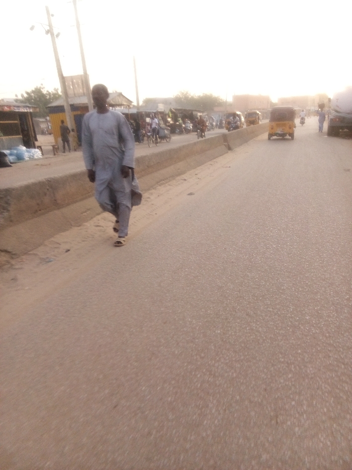The image captures a bustling street scene in what appears to be a semi-urban or urban area, identified by the context as Tudun Murtala, Nigeria. In the foreground, a man in a light gray traditional garment, possibly a kaftan or djellaba, walks along the edge of a paved road towards the left. He is wearing sandals.
The road occupies the center and right side of the frame, showing light wear and a slightly dusty appearance. On the left side of the road, a concrete barrier separates the main thoroughfare from a lower section where shops or stalls are set up. Numerous people are visible in the mid-ground on this left side, some gathered around the stalls, others appearing to be walking or riding motorcycles. Several motorcycles are parked or moving slowly. Further down the road on the right, multiple yellow auto-rickshaws, commonly known as Keke Napep in Nigeria, are visible, suggesting active traffic. A larger vehicle, possibly a truck, is partially seen on the far right. Utility poles line the left side of the road, indicating infrastructure.
The lighting suggests late afternoon or early morning, with a soft, warm, and somewhat hazy glow across the sky and scene. The atmosphere appears calm but active, depicting everyday life in the area. There is no visible text in the image.