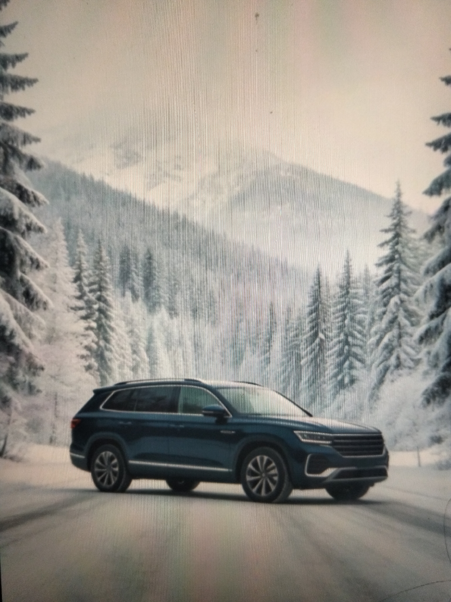 A dark blue SUV is parked on a snow-covered road, surrounded by snow-laden pine trees and a mountain range in the background. The sky is overcast, suggesting a wintery atmosphere. The scene appears to be a promotional image for the vehicle, emphasizing its capability in snowy conditions. There are no people visible in the image. The location is not Payagaboh, Indonesia, as the environment depicted is characteristic of a cold, mountainous region, not a tropical climate.