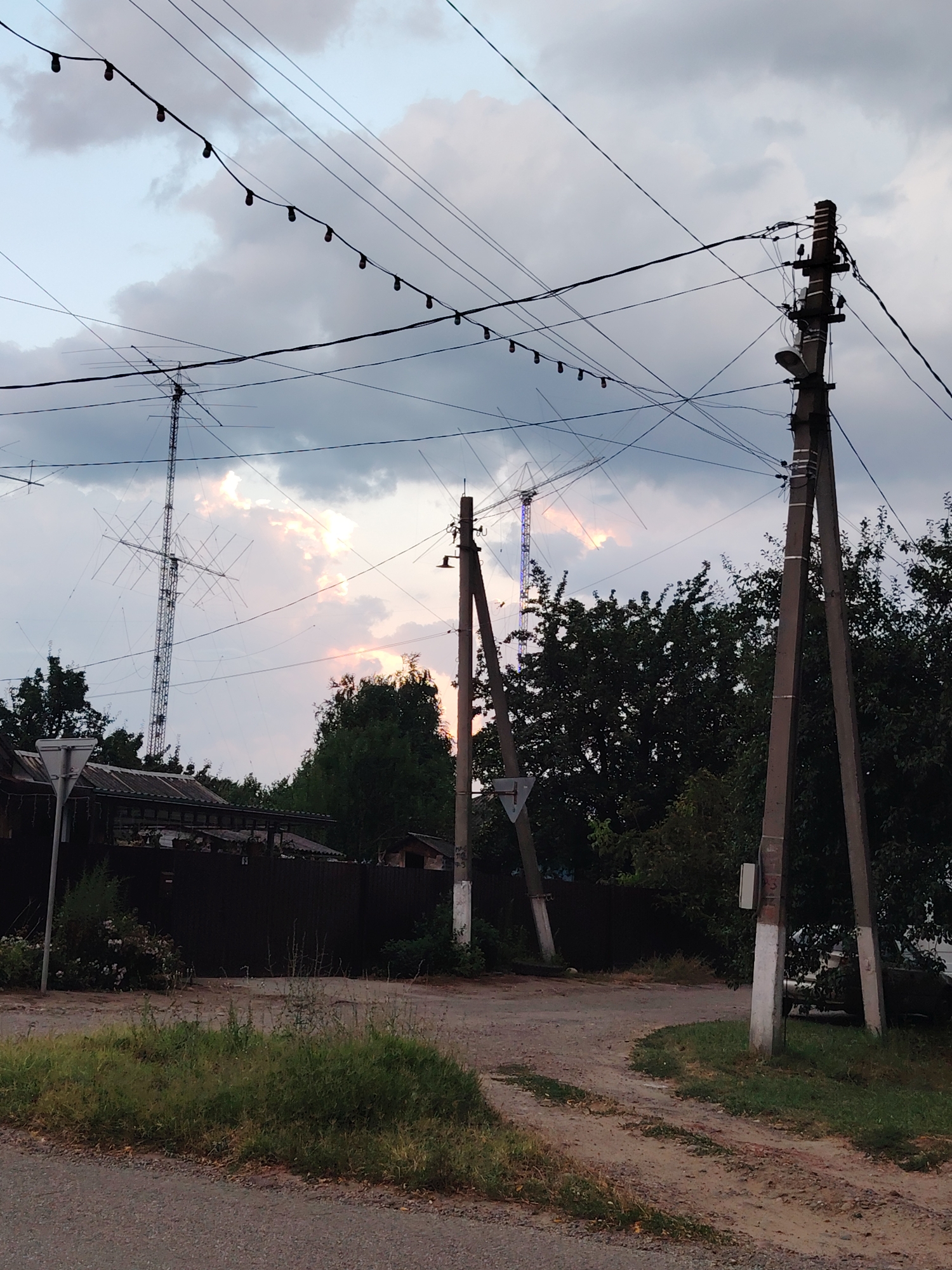 The image depicts an outdoor scene, possibly in Volnoye, Russia, dominated by a network of power lines and utility poles against a cloudy sky. In the background, a tall lattice tower, likely a radio or transmission tower, is visible with a complex antenna structure attached. The sky shows hints of sunset colors breaking through the clouds. In the foreground, a dirt road curves through an area with scattered green vegetation. A dark fence runs along the side of the road, with glimpses of buildings and trees behind it. The overall impression is one of a rural or semi-rural environment with existing infrastructure.