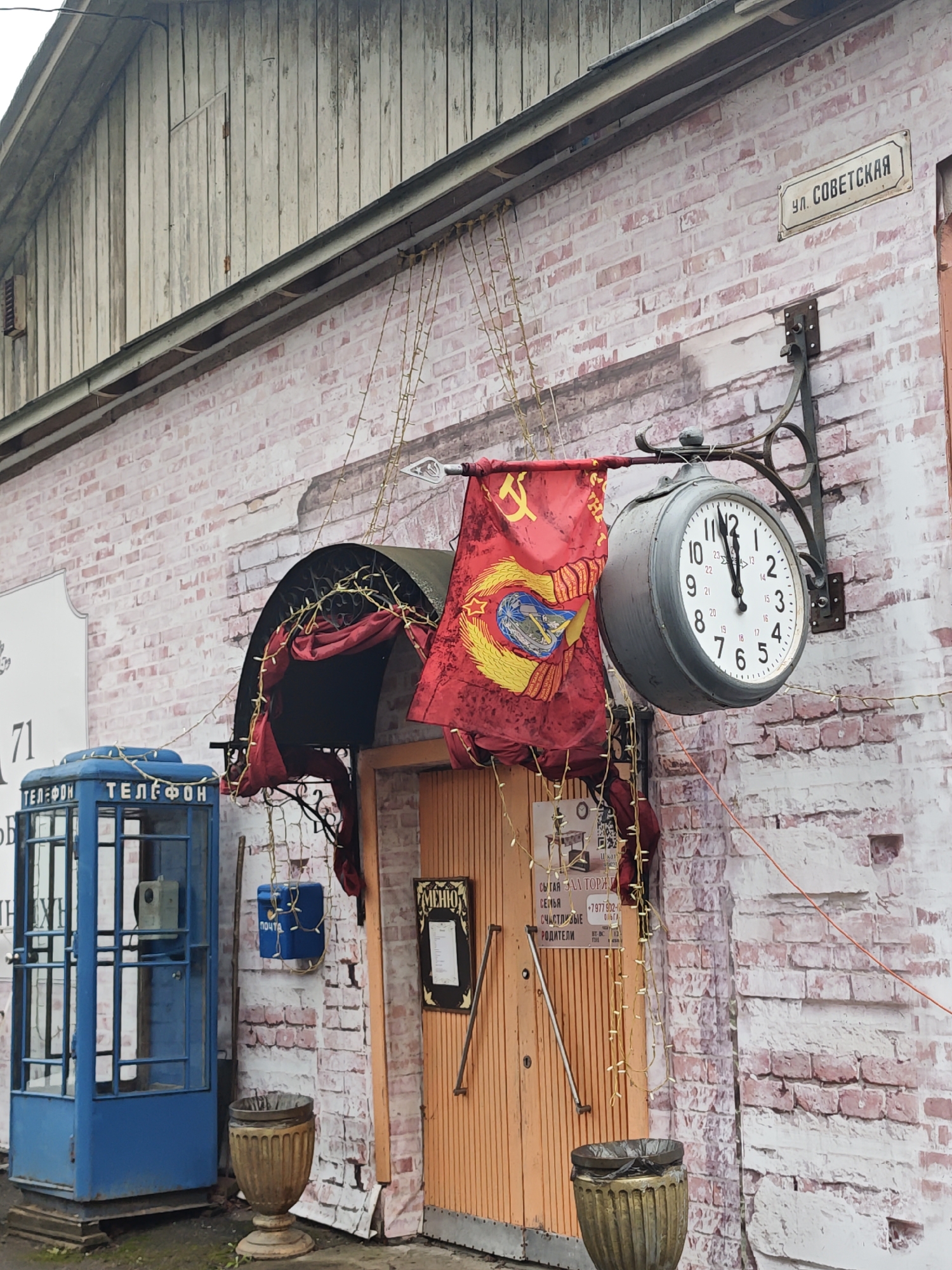 The image captures a Soviet-themed facade in the city of Ольгово, Russia. The building is painted with a faux brick pattern and displays a sign that reads "Ул. Советская", likely indicating "Soviet Street" in Russian. A large, vintage-style clock is mounted on the wall, showing a time of approximately 11:58 or 12:02, hung alongside a red flag adorned with the Soviet hammer and sickle emblem. An awning decorated with red fabric and string lights covers the entrance, and a faded blue telephone booth stands to the left of the door. Below the booth is a decorative urn. Another blue post box can be seen to the right of the telephone booth. Additional signage and decorative elements are present on or near the entrance, adding to the Soviet-era aesthetic. The building's upper section is clad in weathered wooden planks.
