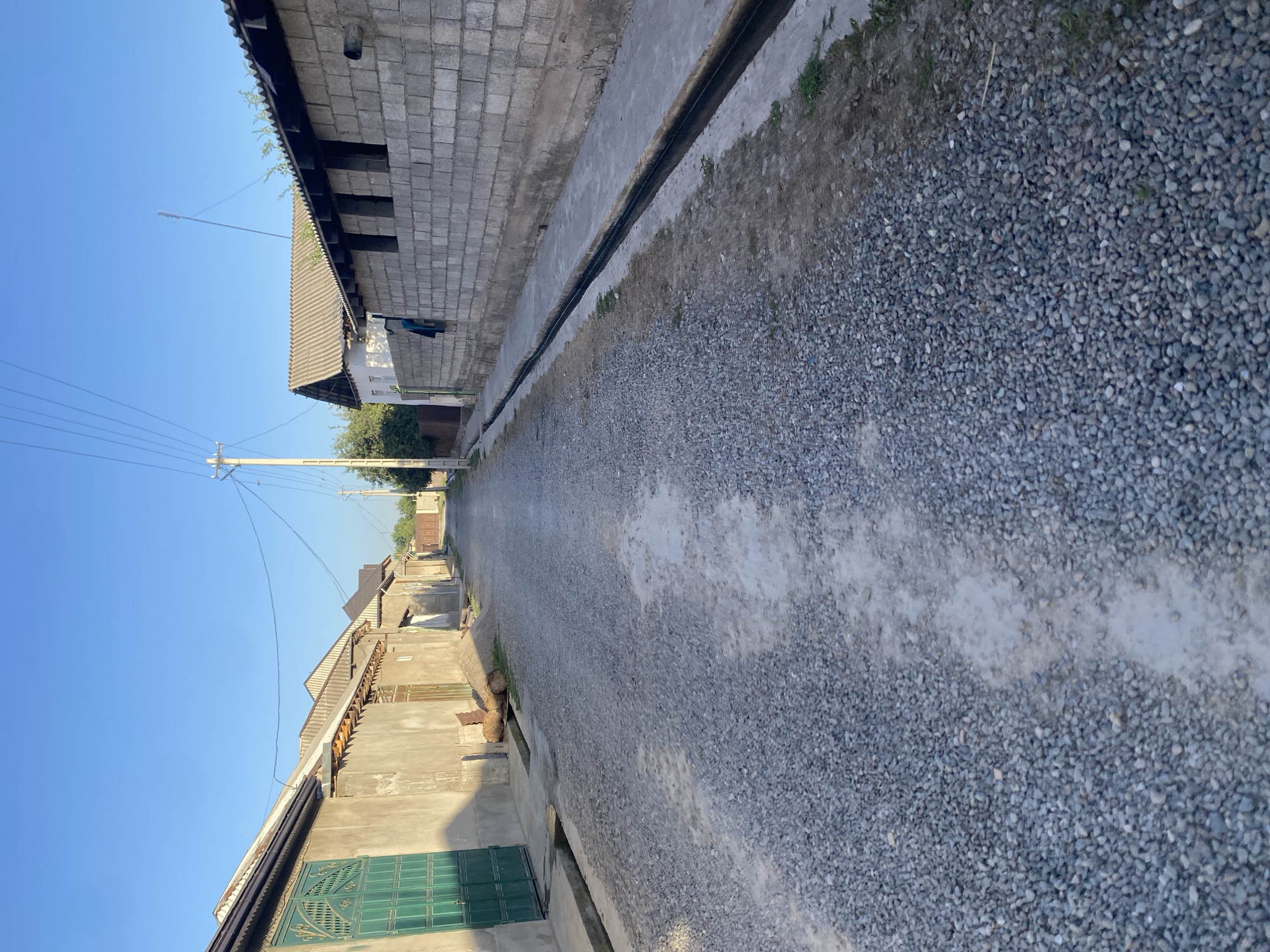 The image shows a gravel road in a residential area, likely in the city of Боғистон, Tajikistan. The road is lined with buildings on both sides. On the left, there are several single-story structures with plastered walls and pitched roofs. One building features a large, ornate green metal gate. On the right, there is a longer, one-story building constructed of grey concrete blocks with an open, roofed section. A dark, narrow drainage ditch runs along the edge of the road in front of this building. Overhead power lines stretch across the clear blue sky. The overall impression is of a somewhat unpaved, rural or semi-urban street.