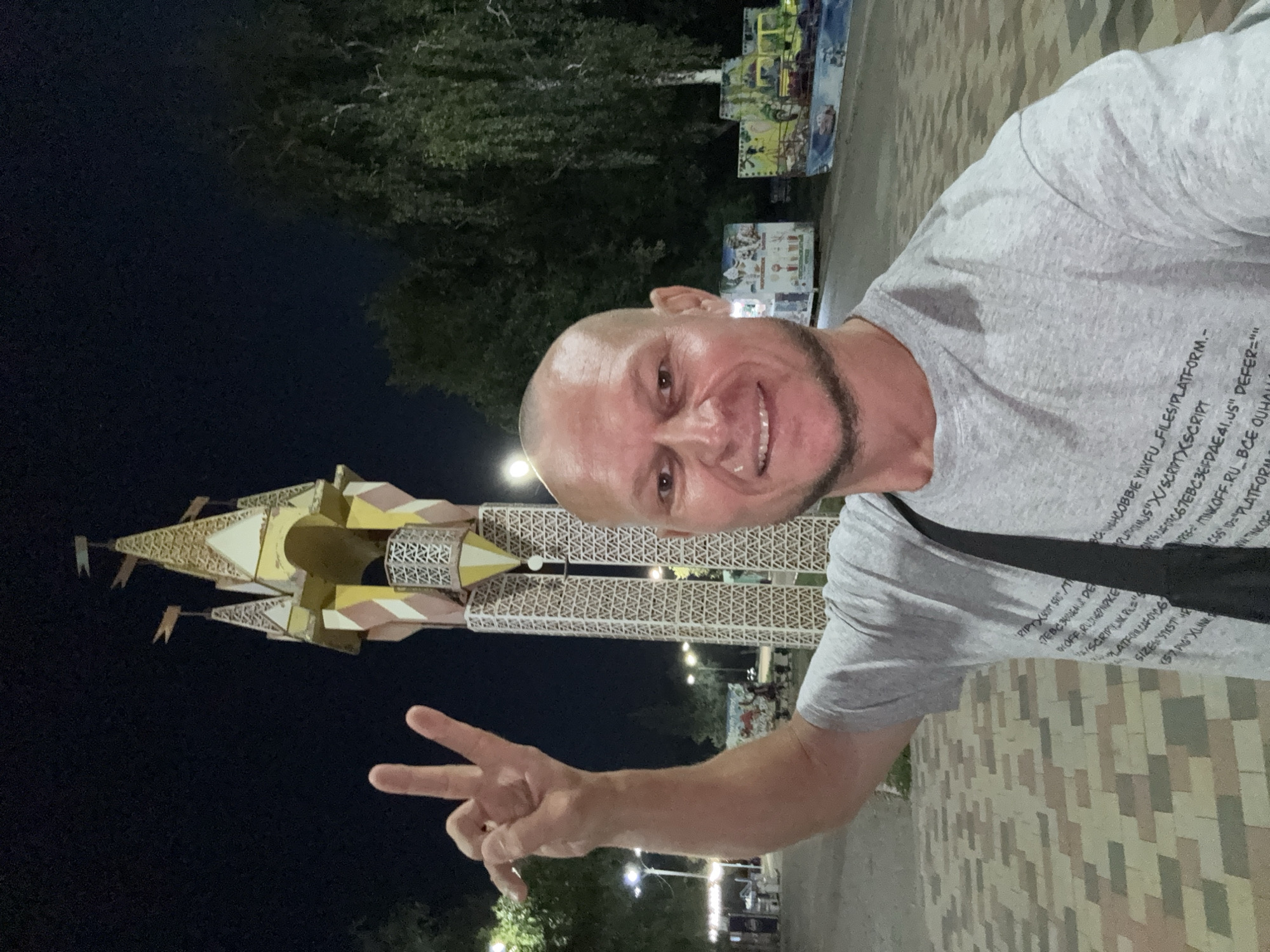 A bald, middle-aged Caucasian man with a goatee is taking a selfie outdoors at night. He is wearing a gray t-shirt with text printed on it and a black strap across his chest. His left arm is raised, and he is making a peace sign with his fingers. Behind him, a decorative tower with a pointed, castle-like structure at the top is visible. The tower is illuminated and appears to be part of an amusement area, with what looks like carnival rides or attractions in the background. The ground is paved with interlocking bricks. Trees are present in the background, and there are streetlights illuminating the scene. The overall atmosphere suggests a nighttime visit to a local park or amusement venue in Oskemen, Kazakhstan.