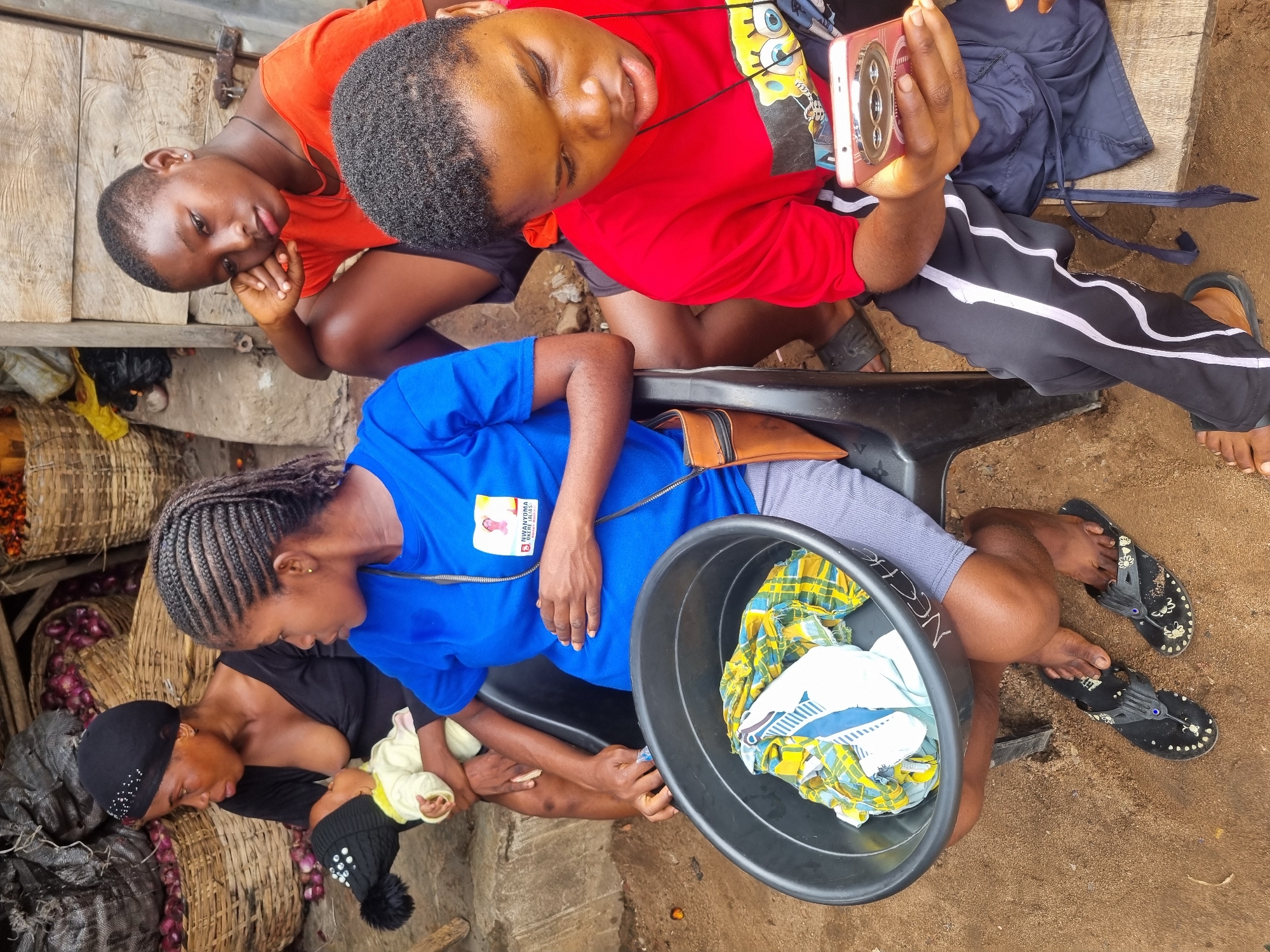 This image captures an intimate outdoor scene in Fegge, Nigeria, likely within a market or street-side area, characterized by baskets of red onions and a dirt ground. The setting suggests a casual, everyday environment.
At the center, a woman in a black top and a black beanie is tenderly cradling and breastfeeding a baby, who is dressed in light yellow clothing and a black beanie with a pompom. Next to her, a woman with braided hair, wearing a bright blue zip-up top, leans forward, appearing to observe the baby and mother with attention. A badge on her shirt is visible, displaying the text "NIFANYENYOMA OKECHUKWU" and a partial phone number. Close to her is a black plastic tub holding an assortment of clothes, including a vibrant yellow and blue patterned fabric.
Further in the frame, another young woman in a red t-shirt, adorned with a Spongebob Squarepants graphic, is intently looking at a pink/rose gold mobile phone with a prominent camera setup. She wears black track pants with white stripes. Behind her, a young woman in an orange t-shirt and dark shorts, with short hair, looks towards the viewer, resting her chin on her hand, displaying a neutral expression. Wooden planks form part of the background structure behind her.
The scene appears to be taking place during the daytime, under natural light. The women are dressed in casual attire, suitable for the local climate. Two pairs of black flip-flops are visible on the ground. The image portrays a moment of community and daily life, with visible human connection and activity.