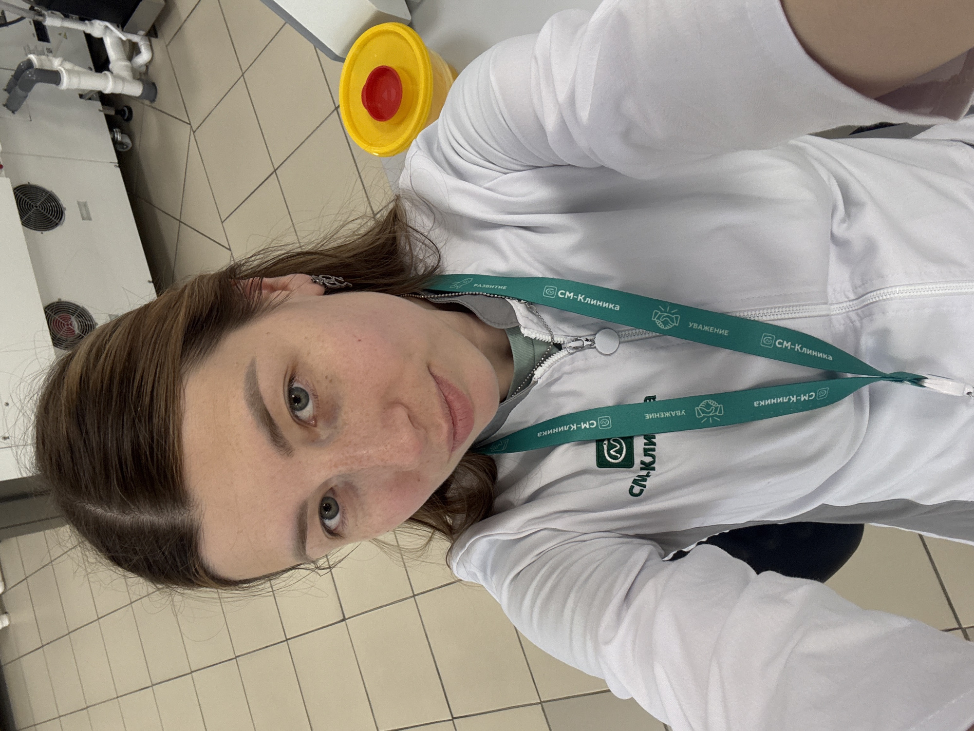 A portrait-style selfie captures a woman indoors, likely within a medical or laboratory setting in Moscow, Russia. The woman, with medium brown hair parted in the middle and light-colored eyes, looks directly at the camera with a neutral to slightly pleasant expression, showing subtle freckles on her face.
She is dressed in a white, zipped medical jacket or lab coat, worn over a light green undershirt. A prominent green lanyard hangs around her neck, featuring the name "СМ-Клиника" (SM-Clinic) in white Cyrillic script, repeated along its length. The lanyard also displays words like "РАЗВИТИЕ" (DEVELOPMENT), accompanied by a circular arrow icon, and "УВАЖЕНИЕ" (RESPECT), next to a handshake icon.
The background shows elements typical of an institutional environment. To the left, white machinery with circular ventilation grilles and visible white piping suggests industrial or medical equipment. On the right, a bright yellow cylindrical bin with a red lid is visible, standing on a light-colored tiled floor. The image appears to be a rotated vertical capture, given the orientation of the woman and the floor tiles. The setting is brightly lit, indicative of artificial indoor lighting.