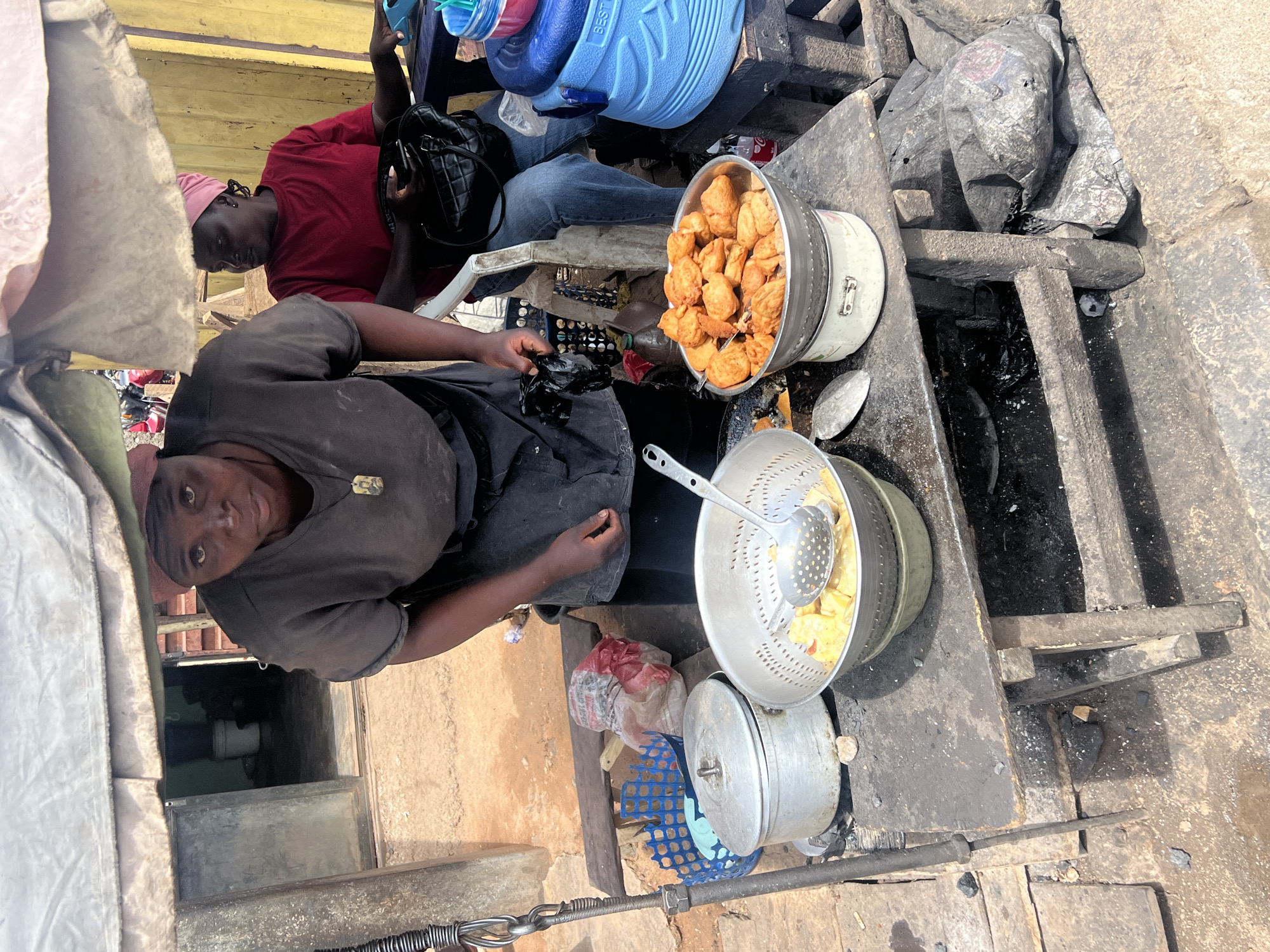 The image shows a food vendor in Gura Top, Nigeria, selling fried snacks outdoors. One woman, wearing a grey shirt and black apron, looks towards the camera as she prepares food. A second woman, wearing a red shirt and pink head covering, sits behind her. A large metal pot of fried snacks sits to the right. Another pot containing ingredients and a slotted spoon is placed below. The cooking area is a dark, worn, wooden structure. Plastic containers are piled on the structure, and plastic bags sit on the ground near the base of the cooking area. The background includes portions of a building and nearby ground surfaces.