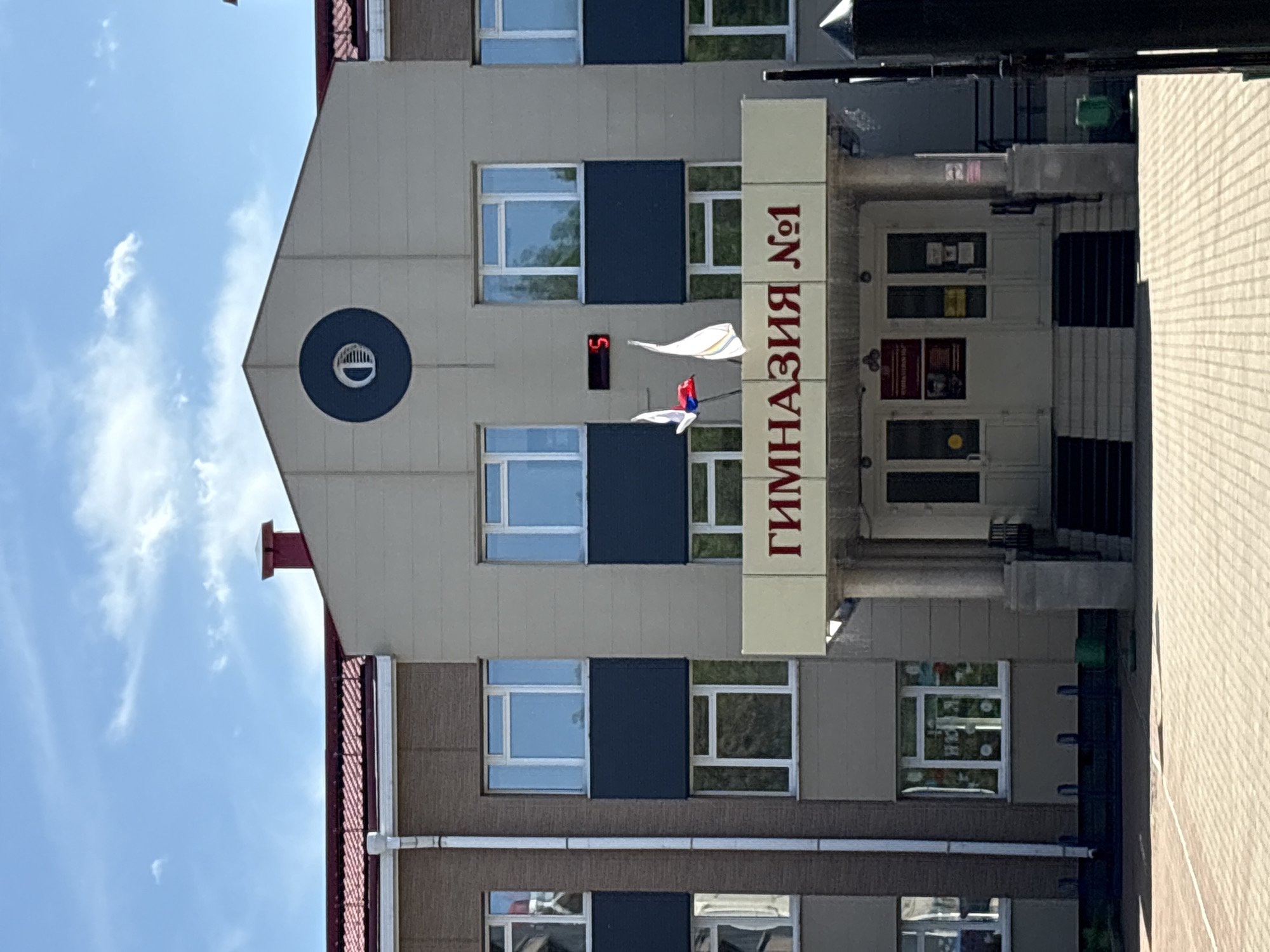 The image shows the exterior of a two-story building, identified by signage as "Гимназия №1" (Gymnasium No. 1). The building features a light-colored facade with rows of windows. The central portion of the building has a triangular pediment with a circular dark blue emblem. Above the entrance, a sign in red letters reads "Гимназия №1". Russian flags are visible near the entrance, along with a digital clock displaying "5". The building is set against a bright blue sky with scattered white clouds. The foreground shows a paved area, likely a courtyard or walkway, with metal railings and a green trash can. The architecture suggests a public institution, possibly a school or administrative building, located in the city of Birobidzhan, Russia.