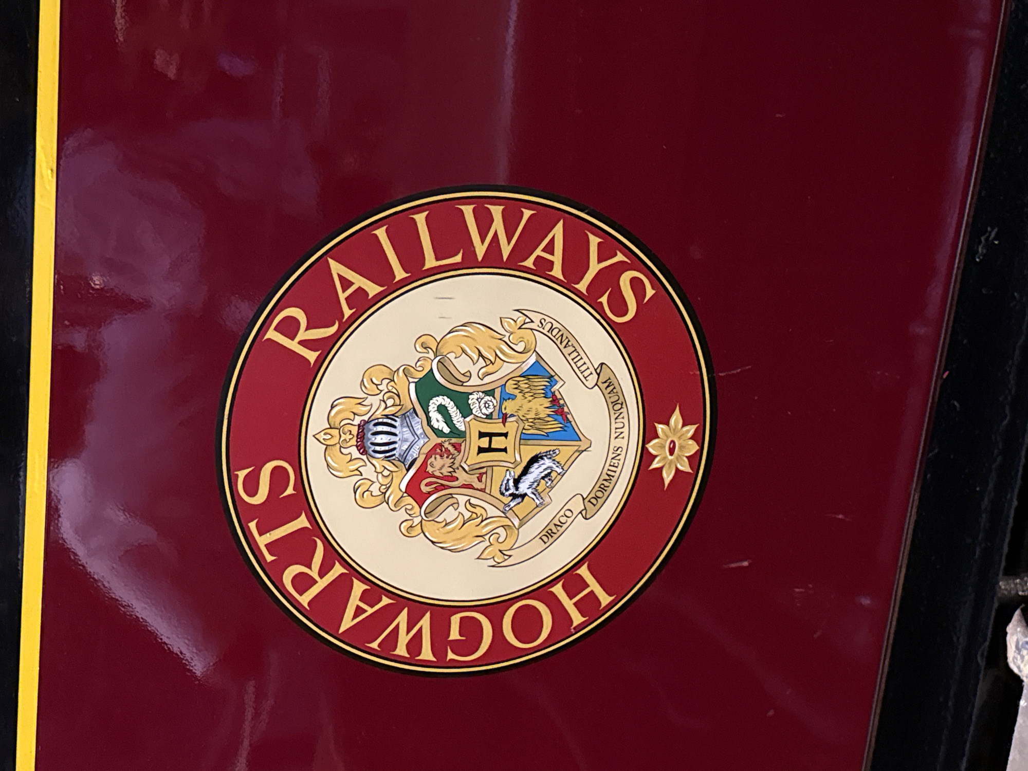 The image shows a close-up of a portion of a maroon-colored train car, likely the Hogwarts Express, given the prominent emblem. The emblem, circular in shape, features the words "HOGWARTS RAILWAYS" in gold lettering around the outer edge. Inside the circle is a detailed crest, divided into four sections, each representing a Hogwarts house: Gryffindor (lion), Hufflepuff (badger), Ravenclaw (eagle), and Slytherin (serpent). A helmet and banner with the Latin motto "DR A C O DORM IE N S N U N Q U AM" are also visible. The train car has a glossy finish, reflecting light and showing some minor scuffs and scratches. A thin yellow line runs horizontally across the top of the train car, and a black metal structure is visible at the bottom. This train is located at King's Cross Station in London, United Kingdom, which is the departure point for the fictional Hogwarts Express in the Harry Potter series.