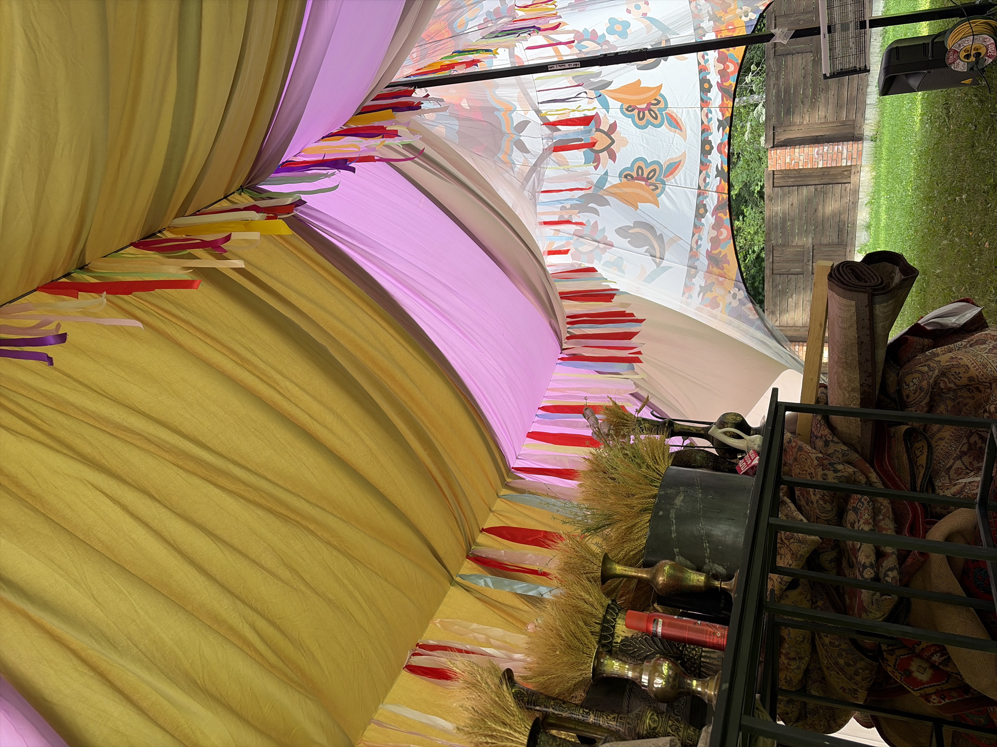 This image depicts the interior of a tent with fabric draped to form a canopy in hues of yellow and pink. Colorful ribbons are strung along the fabric, adding a festive atmosphere. In the foreground, a shelf holds decorative vases and bundles of dried wheat. Below the shelf, rolled carpets and burlap sacks are stacked. The tent's fabric features a patterned design with floral motifs in shades of orange, blue, and brown. Through an opening in the tent, a wooden fence and greenery are visible, suggesting an outdoor setting. The environment appears to be a grassy area, with a speaker and a heating unit situated near the tent. The location is identified as Shebanovo, Russia.