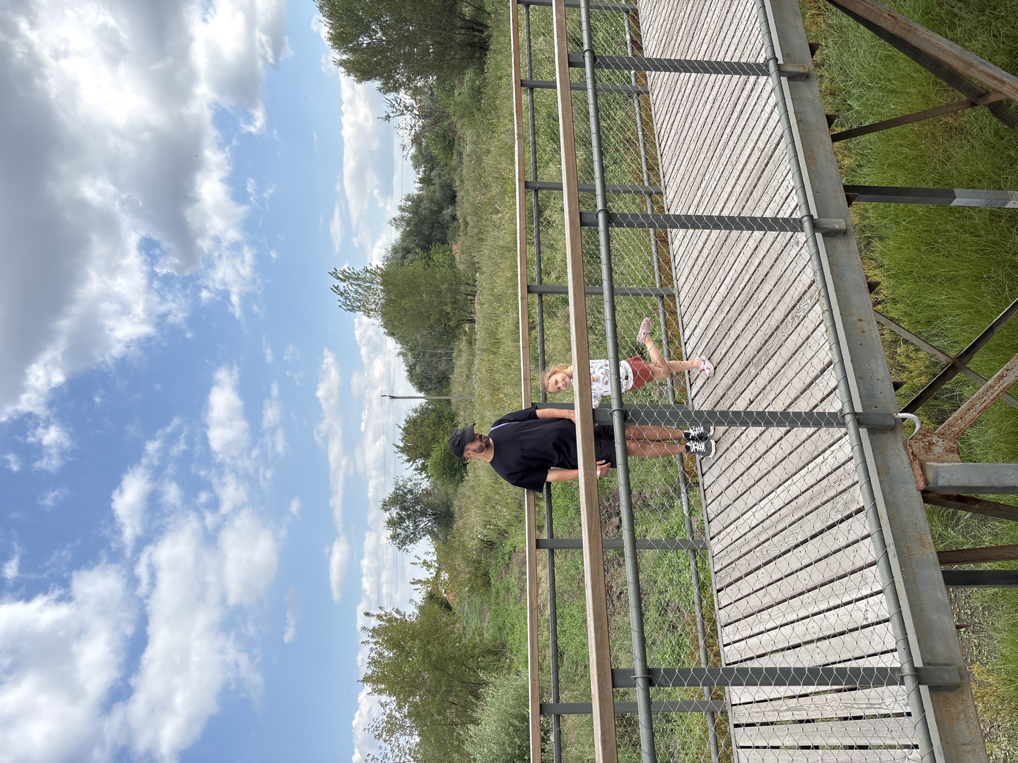 The image shows a father and daughter standing on a wooden footbridge. The father, wearing a black t-shirt, shorts, and a black baseball cap, is standing on the left side of the bridge. The daughter, wearing a white patterned t-shirt and a coral-colored skirt, is standing on the right side of the bridge. The bridge has metal railings with wire mesh infill and wooden planks for the walkway. Behind the bridge, there is a natural landscape with green grass, trees, and a partly cloudy blue sky. The image appears to be taken in an outdoor recreational area, possibly a park or nature reserve. This location is not specified to be in Orenburg, Russia.