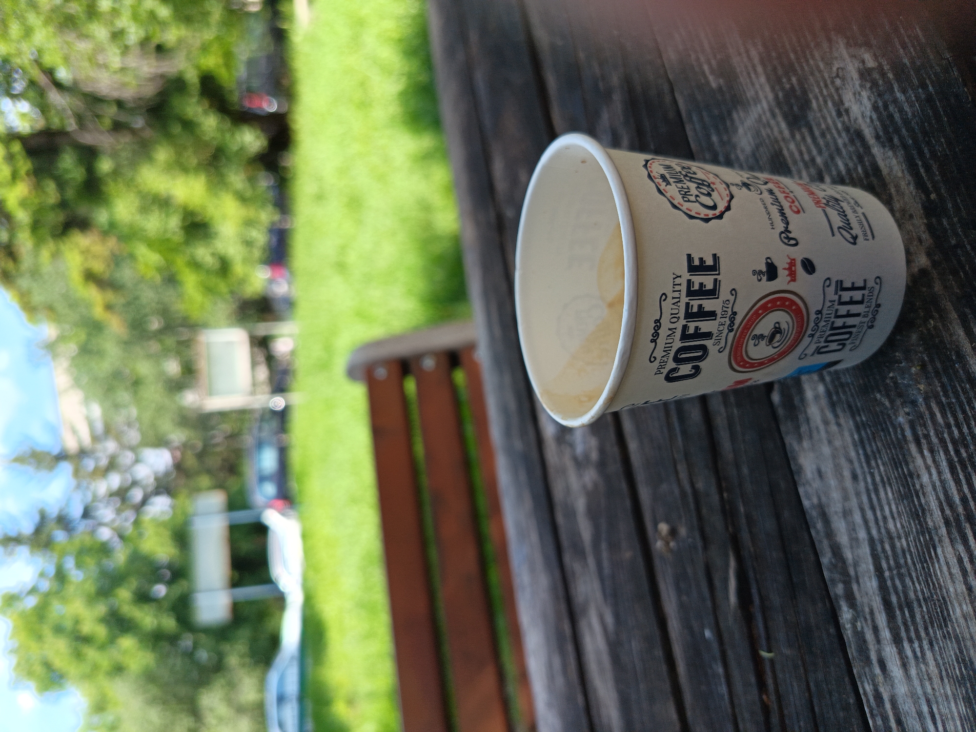 The image shows a paper coffee cup sitting on a wooden bench. The cup is mostly empty, with some coffee residue visible inside. The cup has coffee-related branding on its exterior. The bench is made of weathered wood and has several slats. Behind the bench, there is green grass and trees. The scene appears to be outdoors, possibly in a park. While Sofia, Bulgaria is noted, there is no direct evidence in the image itself to definitively confirm the location.